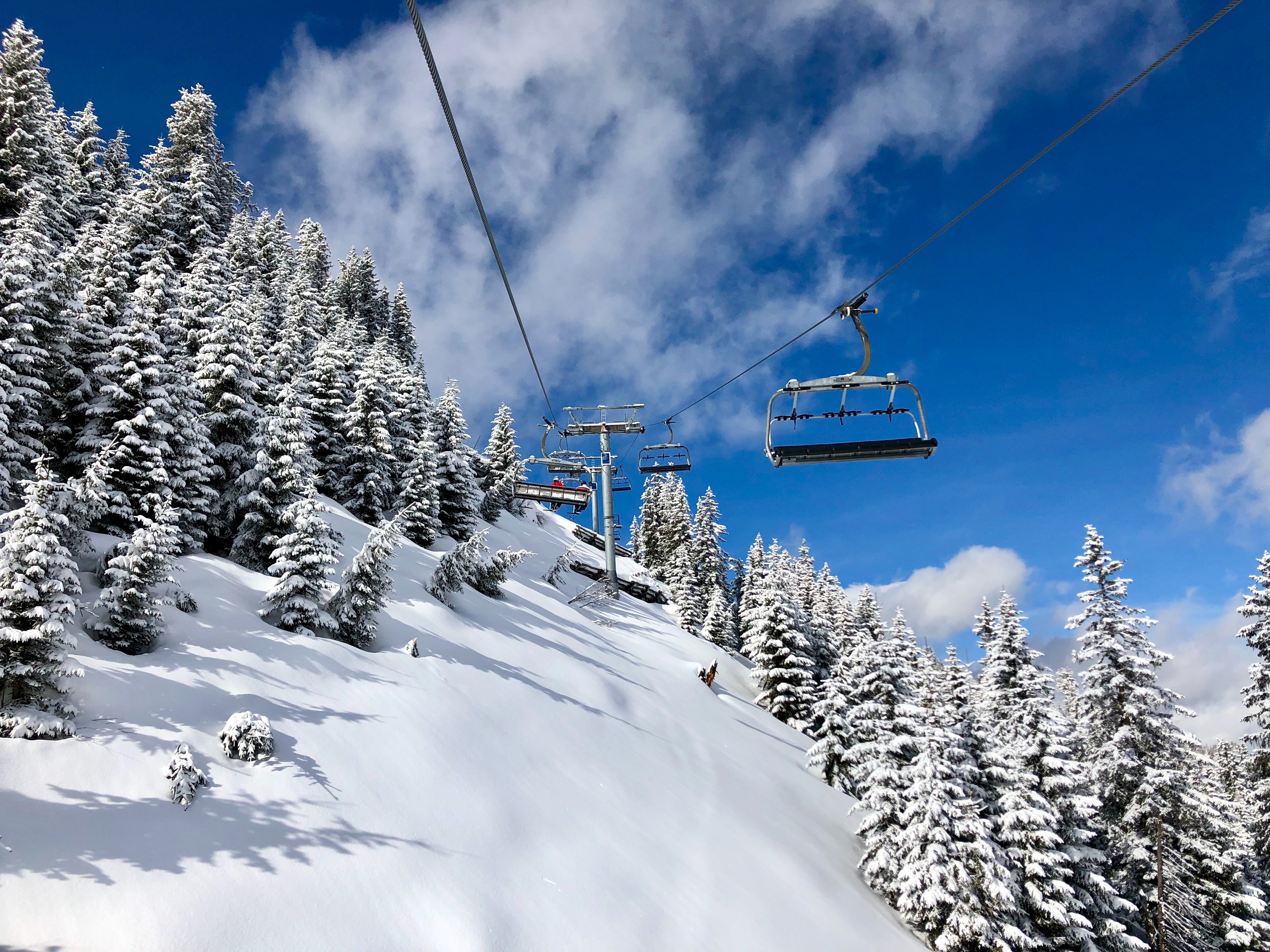 A ski lift with empty chairs ascending a snowy mountain slope lined with evergreen trees under a blue sky with scattered clouds.