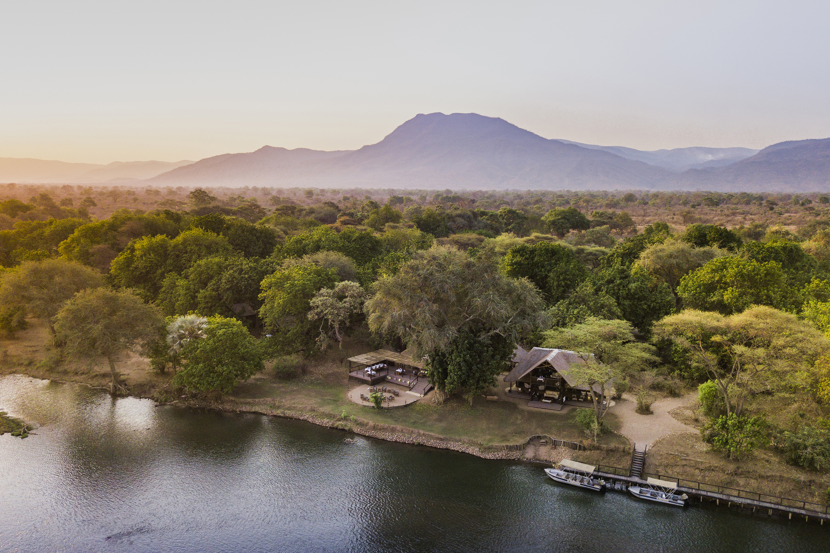 An exterior aerial view of Chiawa Camp's thatched buildings surrounded by trees and backed by mountains beneath a colourful sky at sunset