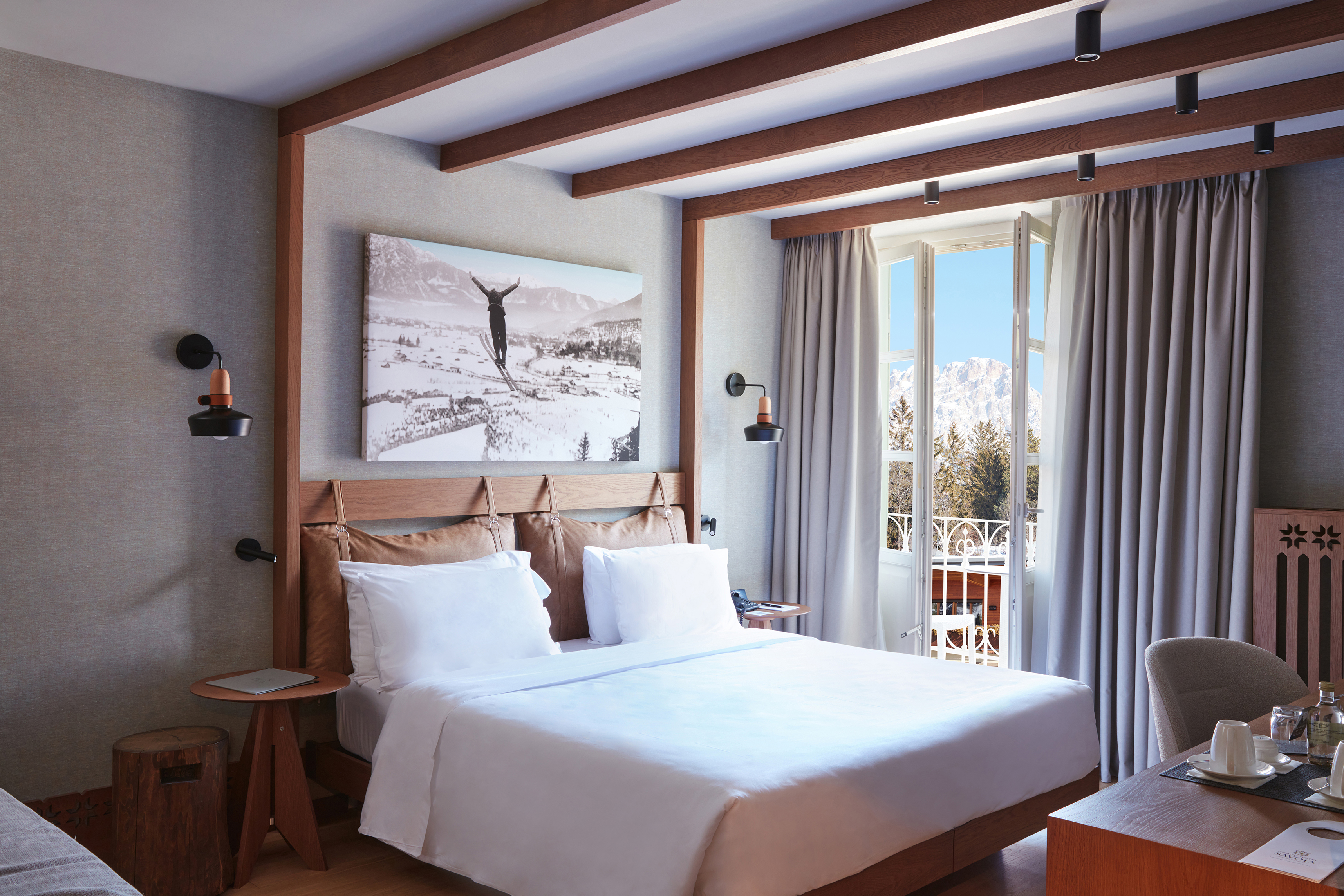 A Premium Collection Room at Grand Hotel Savoia featuring a bed, desk area and skiing artwork with large windows looking out on a terrace to the mountains