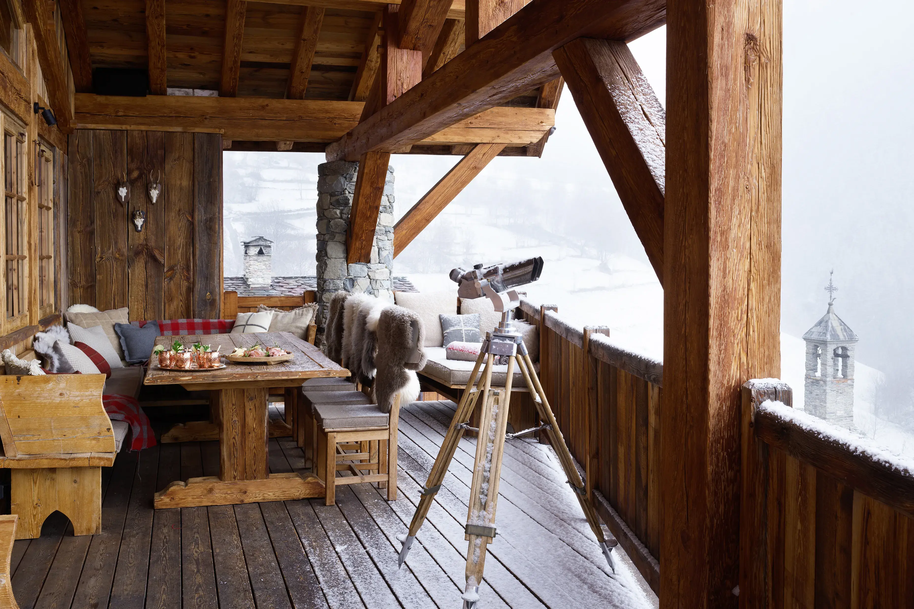 Sun terrace of Chalet Hibou featuring a large dining table set with drinks and snacks and cosy fur blankets, with a telescope pointed out towards a snowy landscape