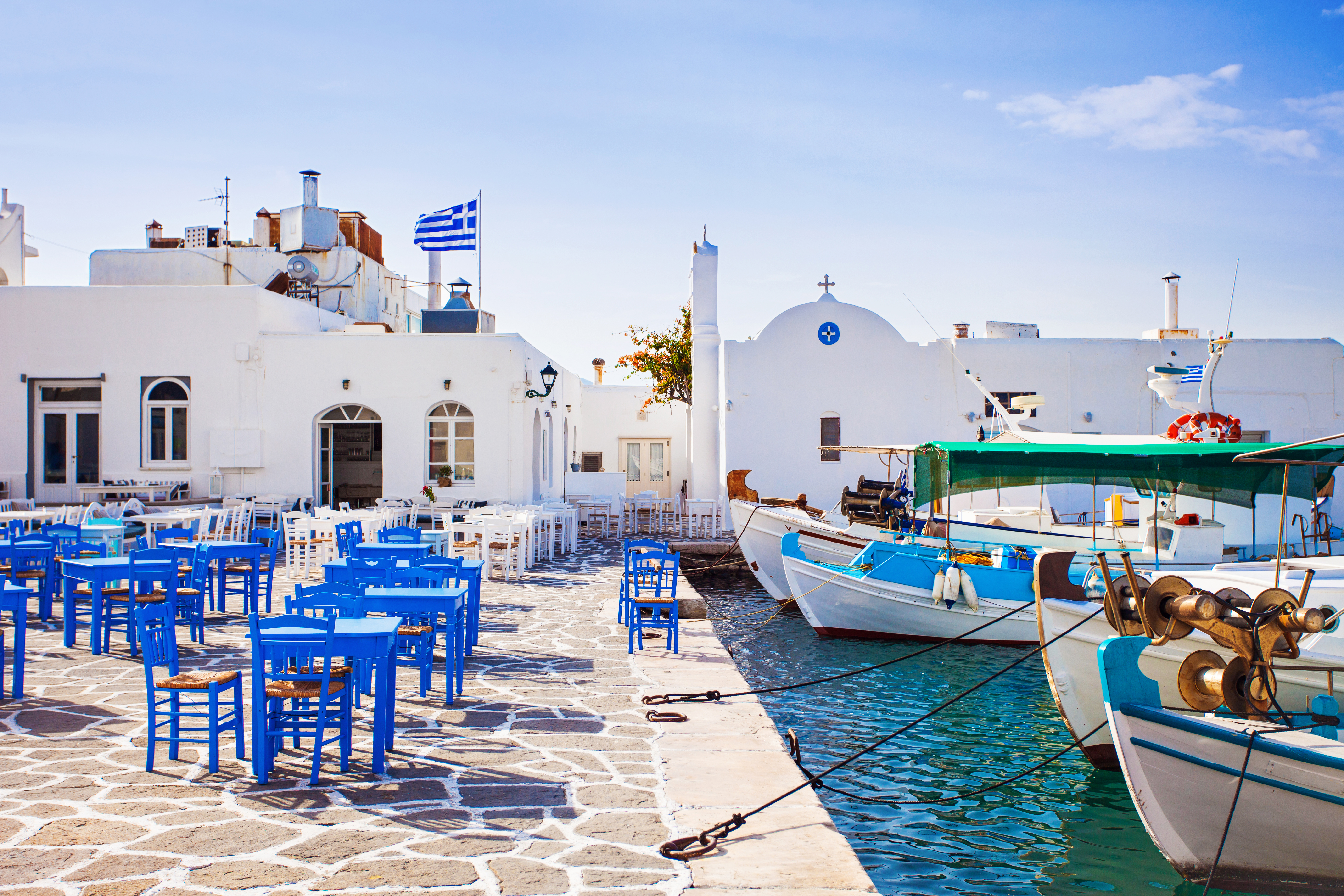 What-washed buildings and bright blue tables and chairs sit next to a marina where small fishing boats are tied up