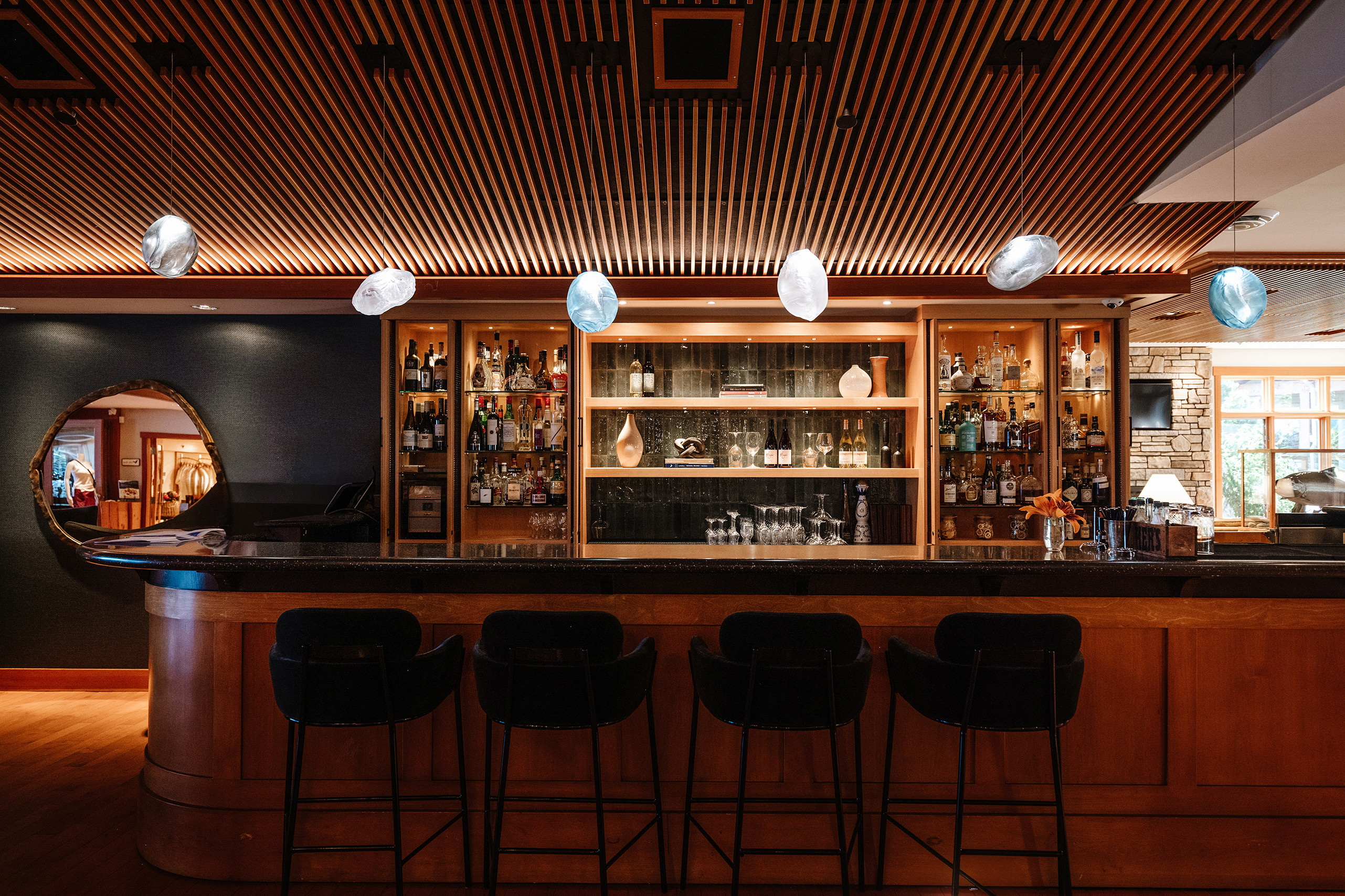 A moody bar with four stools and pebble-shaped light shades in front of a wall filled with bottles and a circular mirror to the side