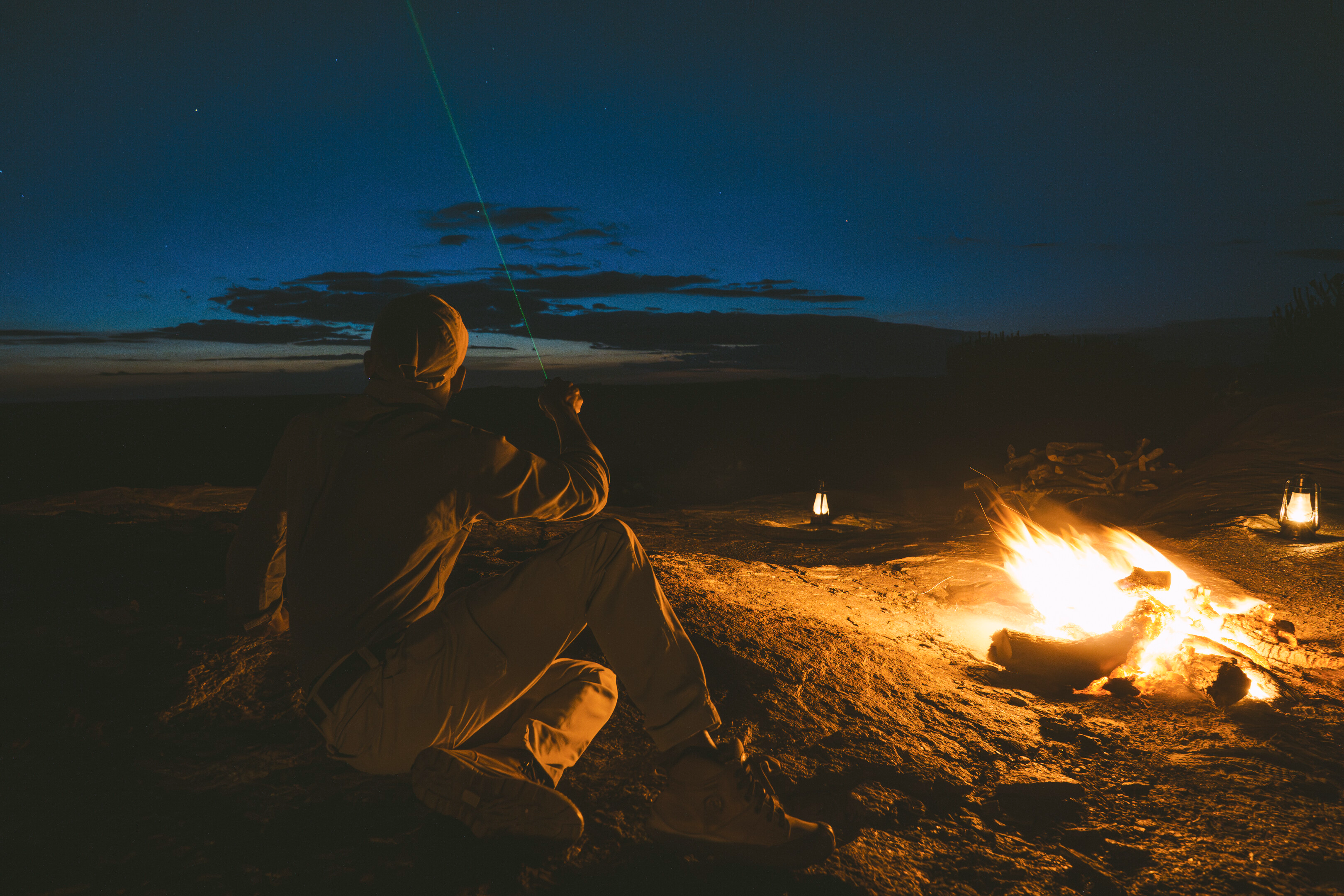 Person seated beside a campfire at night under a star-filled sky, pointing a light towards the horizon.