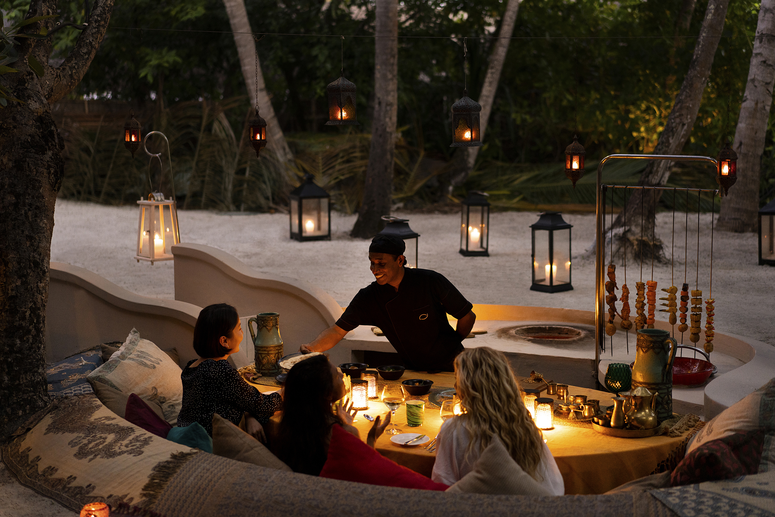 A waiter and three guests sat around a table lit by candles with a glimpse of the beach and larger candles behind