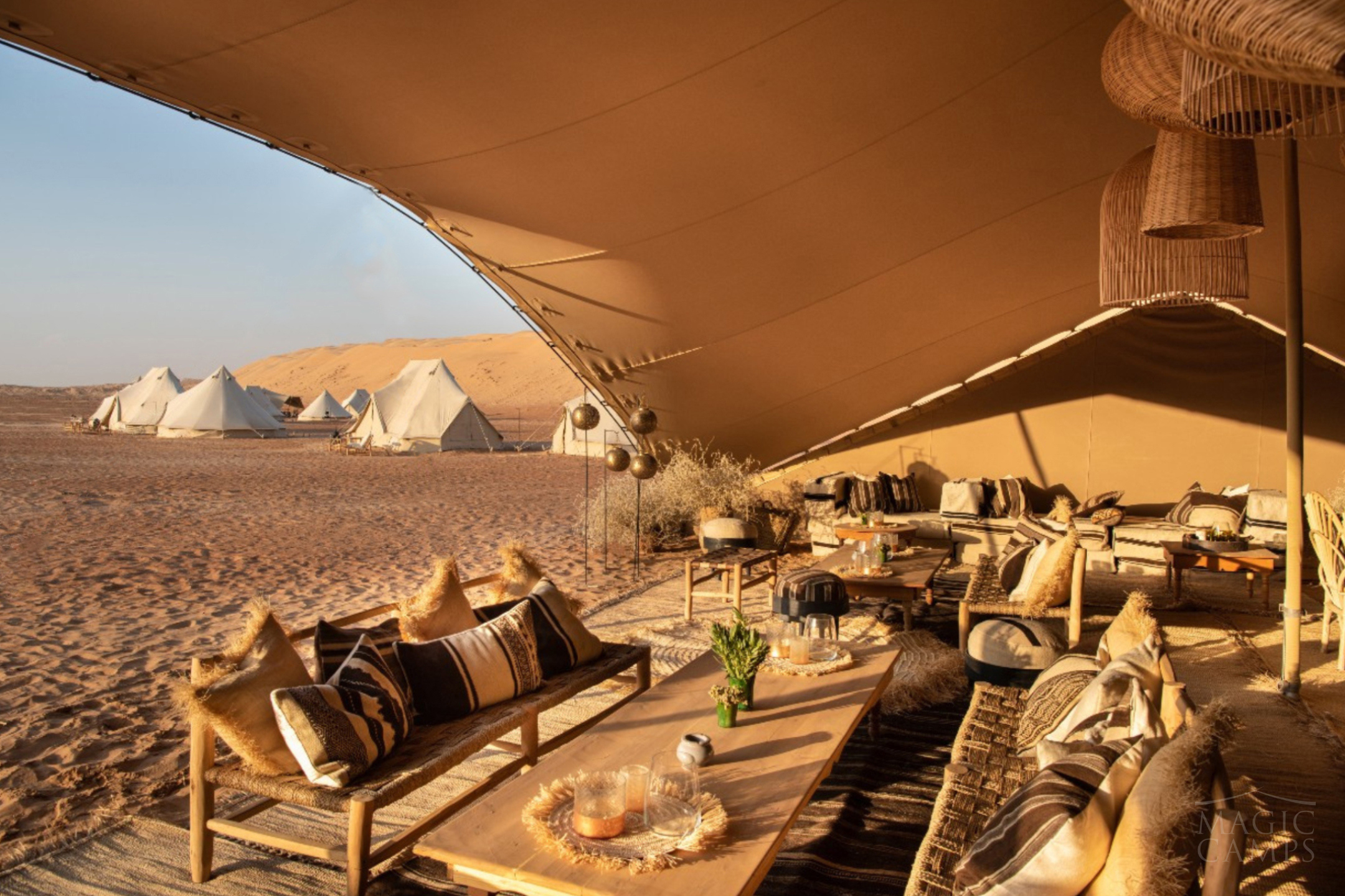 A large, cosy luxury tent featuring wooden chairs and soft furnishing with a view of other tents in the desert outside