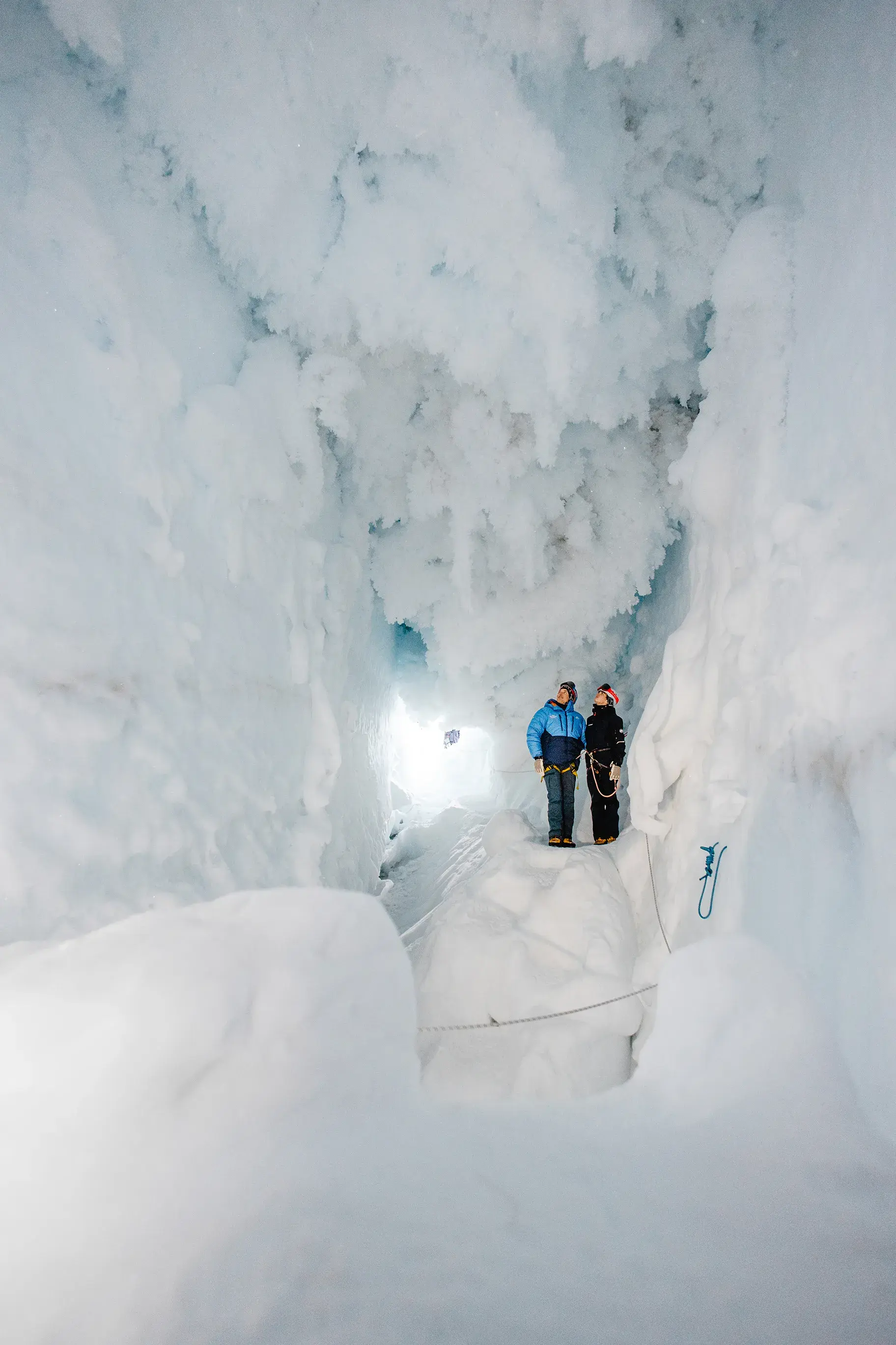 Two people exploring a deep blue‑white ice cave in Antarctica with towering frozen walls and bright light at the entrance.