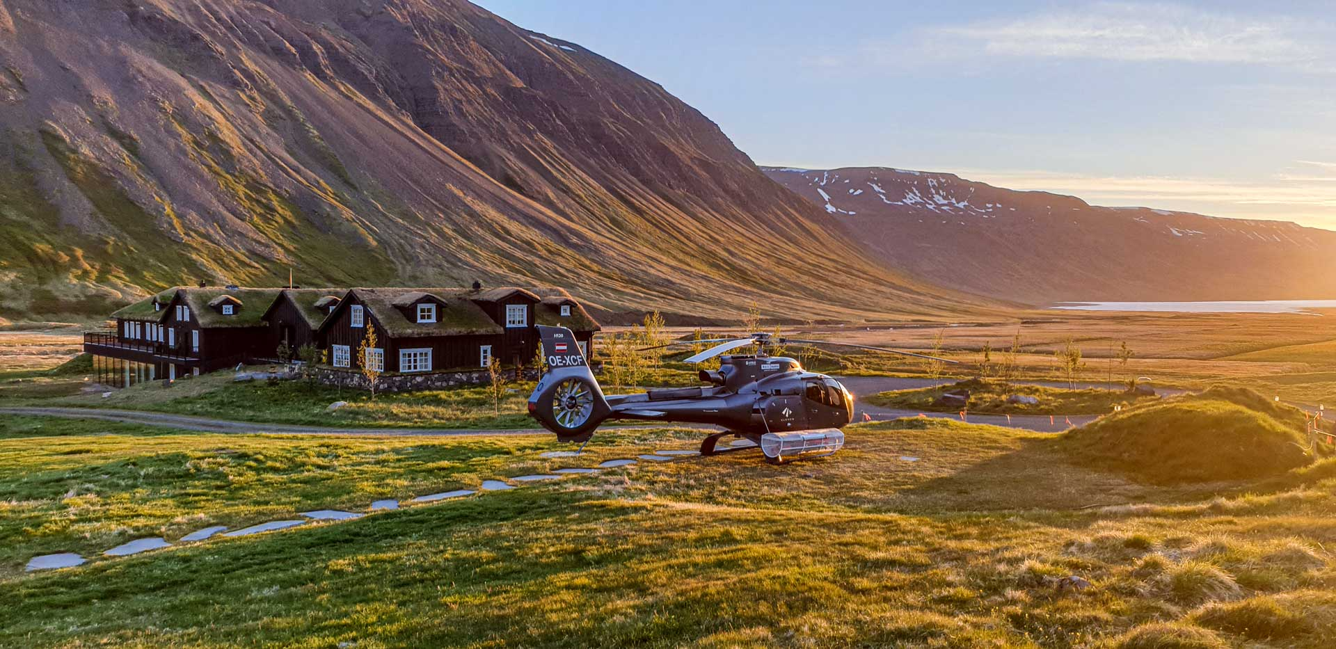Helicopter parked beside a turf‑roof lodge in a wide alpine valley bathed in golden evening light.