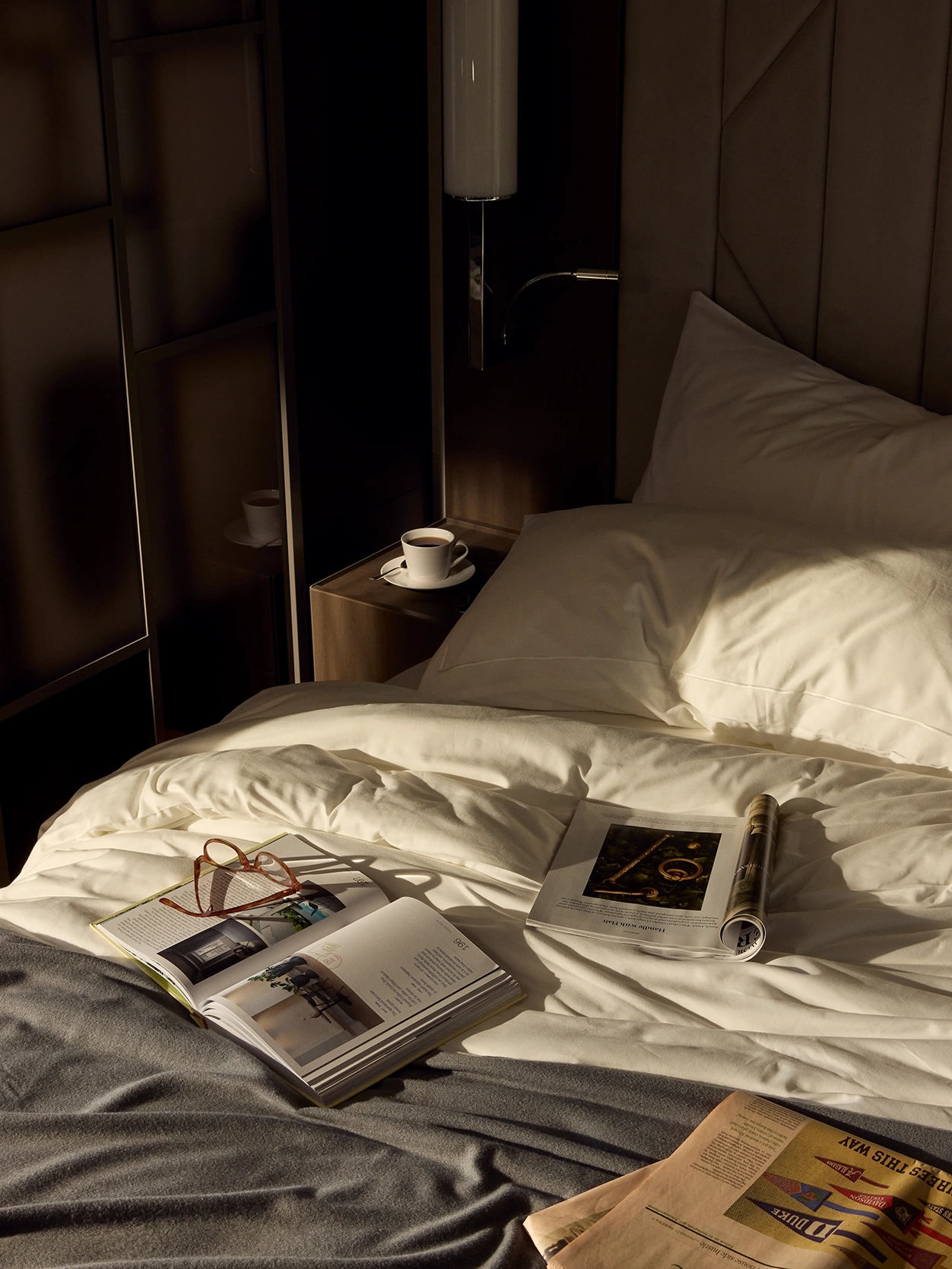 Two magazines and a newspaper left open on an unmade bed with a pair of glasses and coffee in the premier penthouse suite on Explora I cruise ship