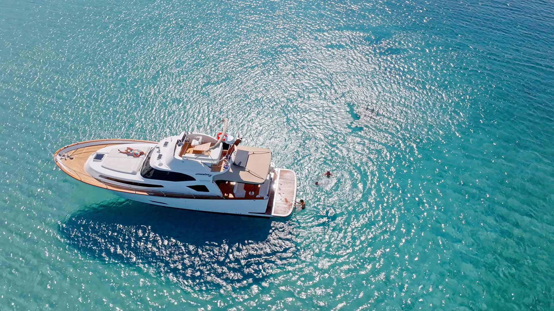 Aerial view of a luxury yacht anchored in clear turquoise water with swimmers nearby.