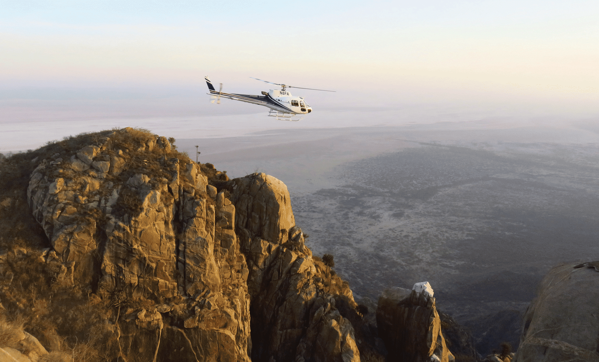 Helicopter flying above rugged rock formations and open savannah landscape in warm golden light.