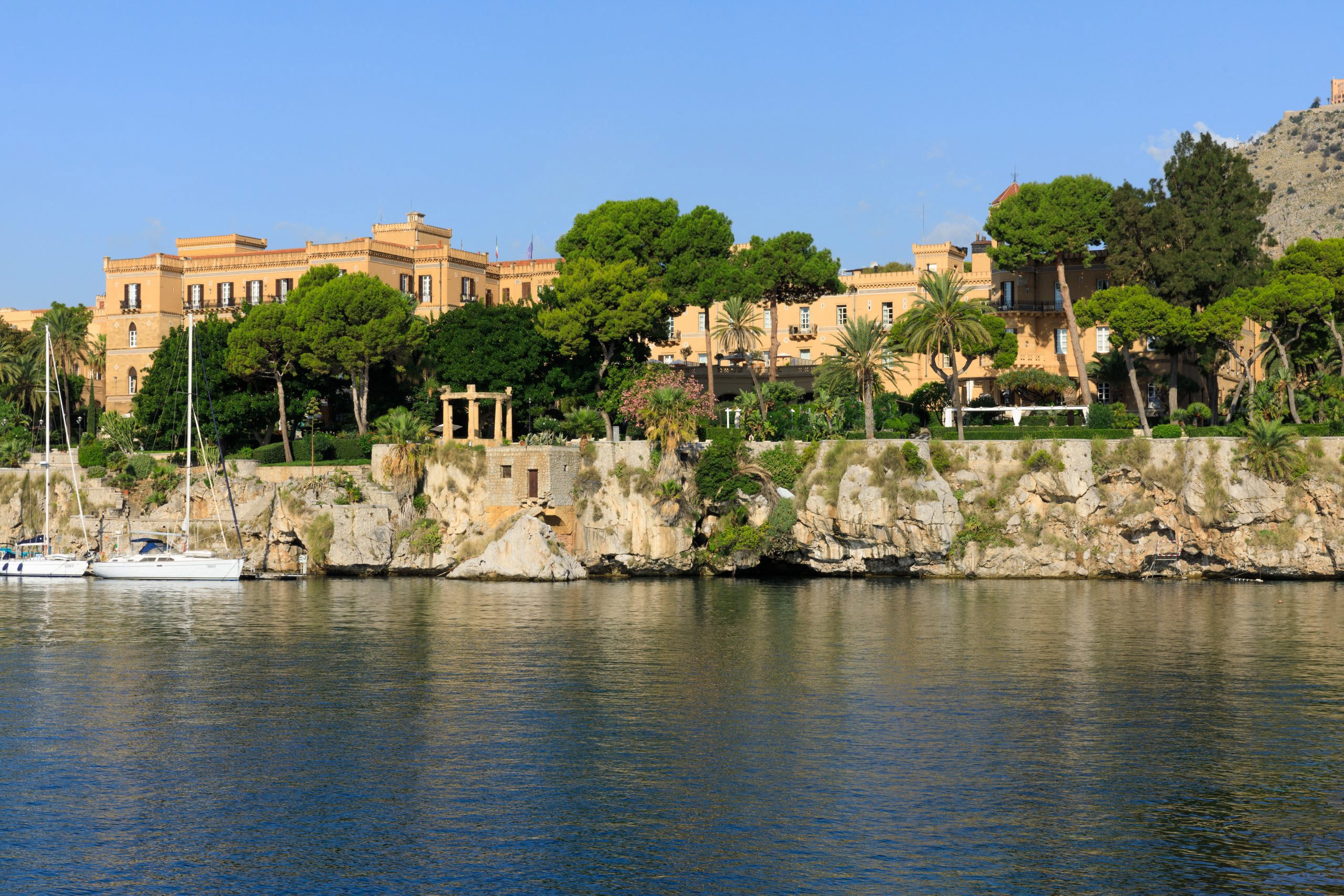 The palatial yellow building of Villa Igiea among trees perched on a rocky coastline with yachts moored in the water below