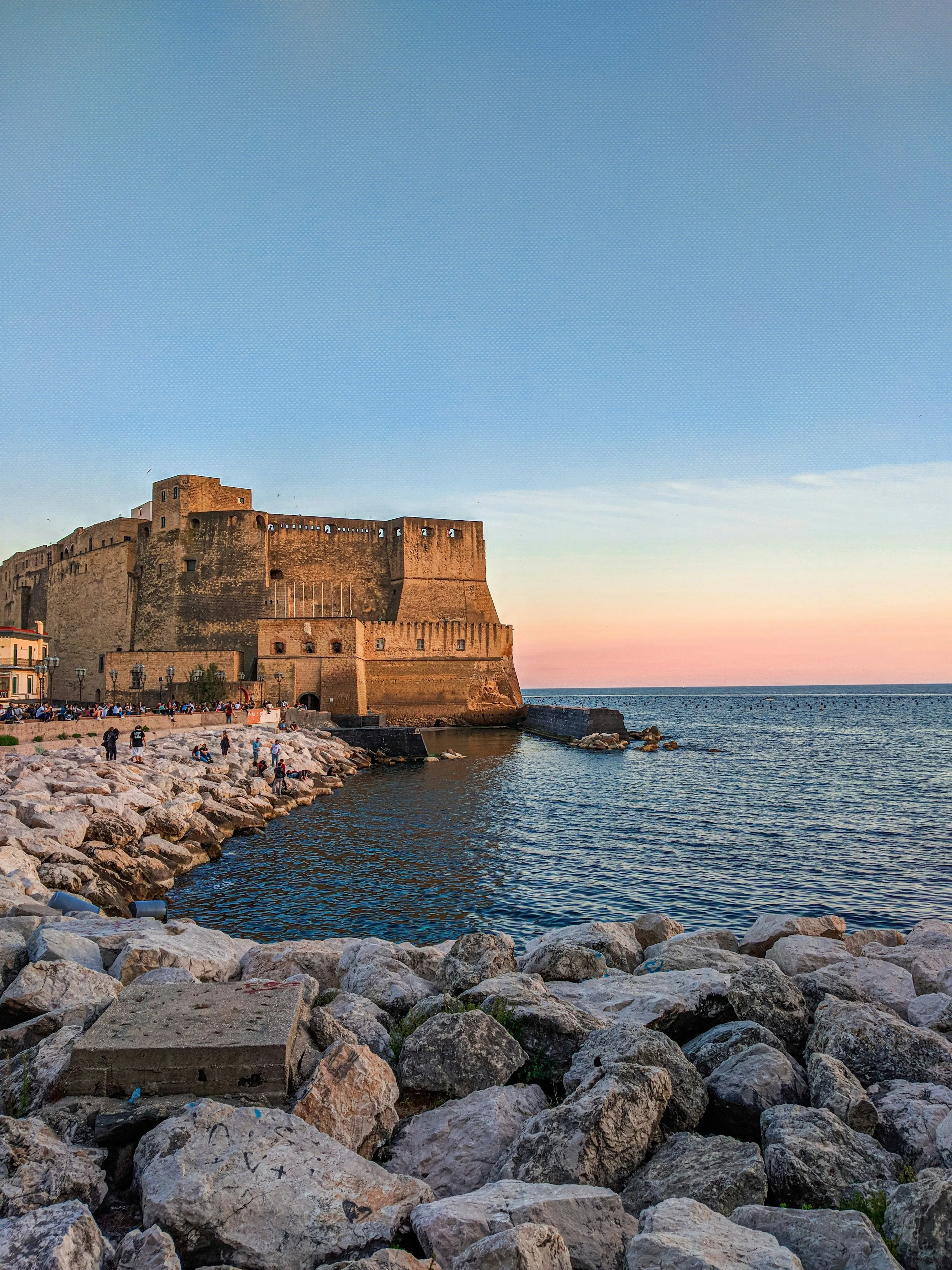 Historic Castel dell’Ovo on the Naples waterfront at sunset with rocky shoreline and calm sea.