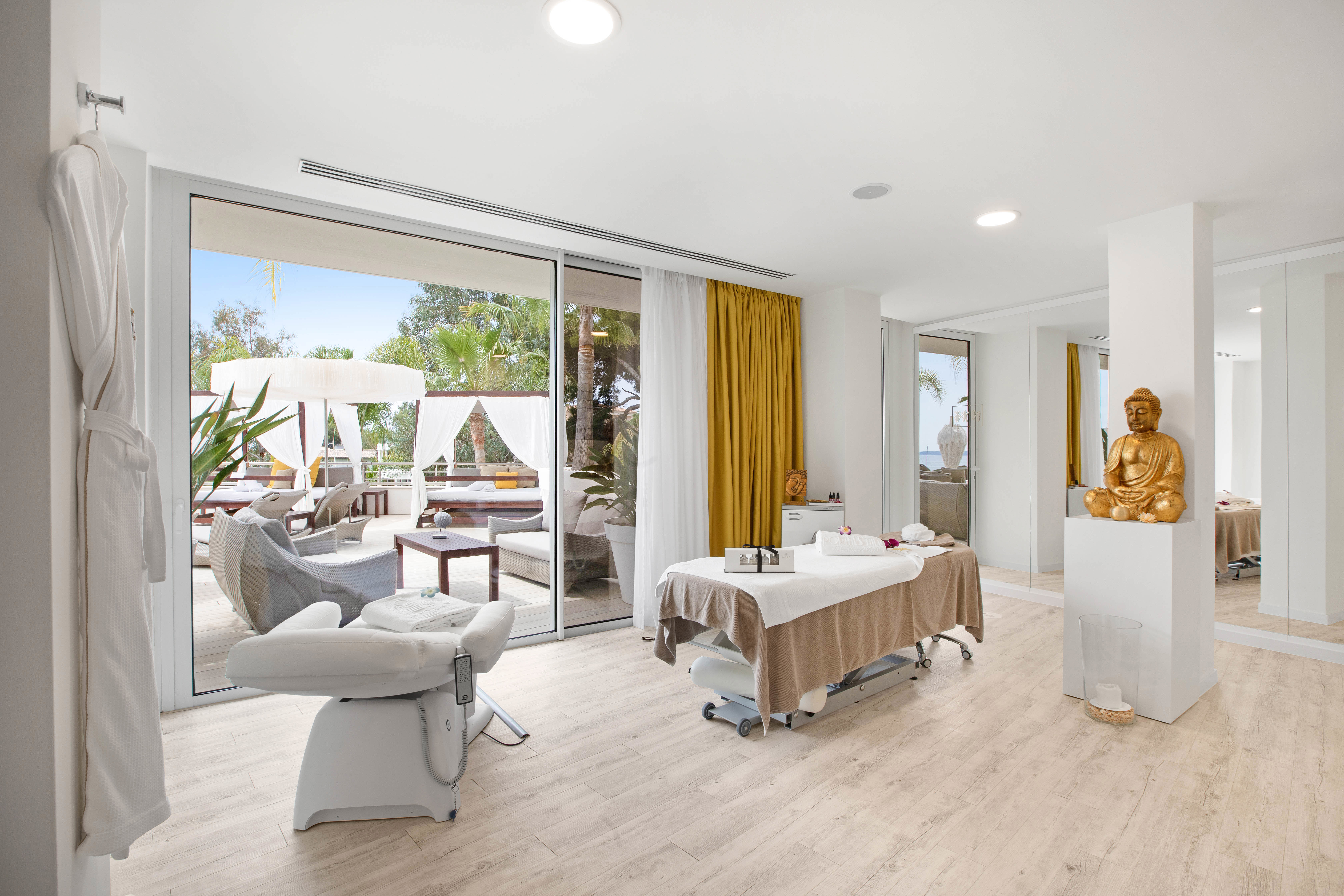 Luxurious spa room at Portals Hills in Mallorca with massage tables, a golden Buddha statue, and views of outdoor cabanas and greenery.