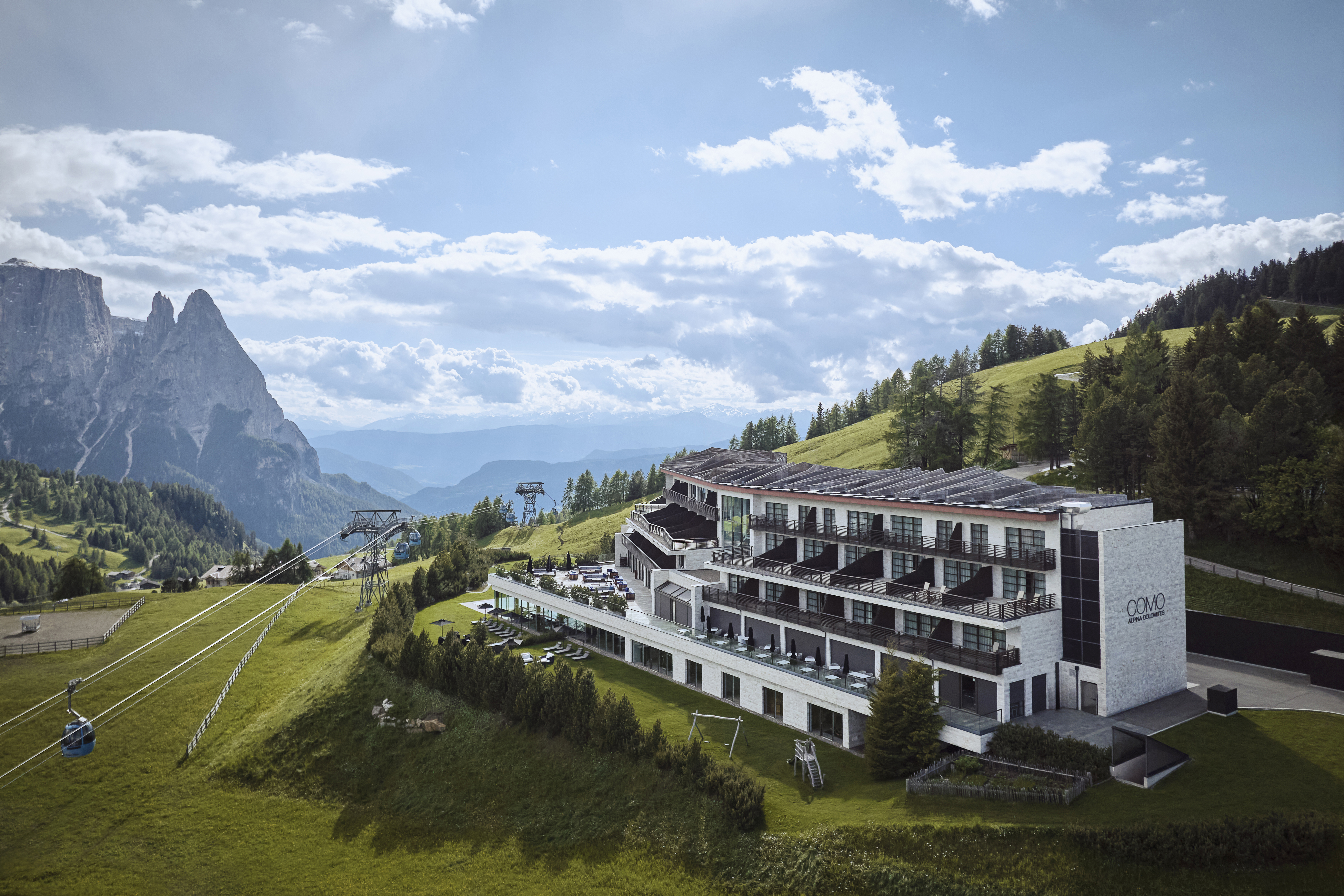 A modern mountain hotel with large windows and a terrace overlooks a snowy landscape during twilight, with rugged peaks in the background.