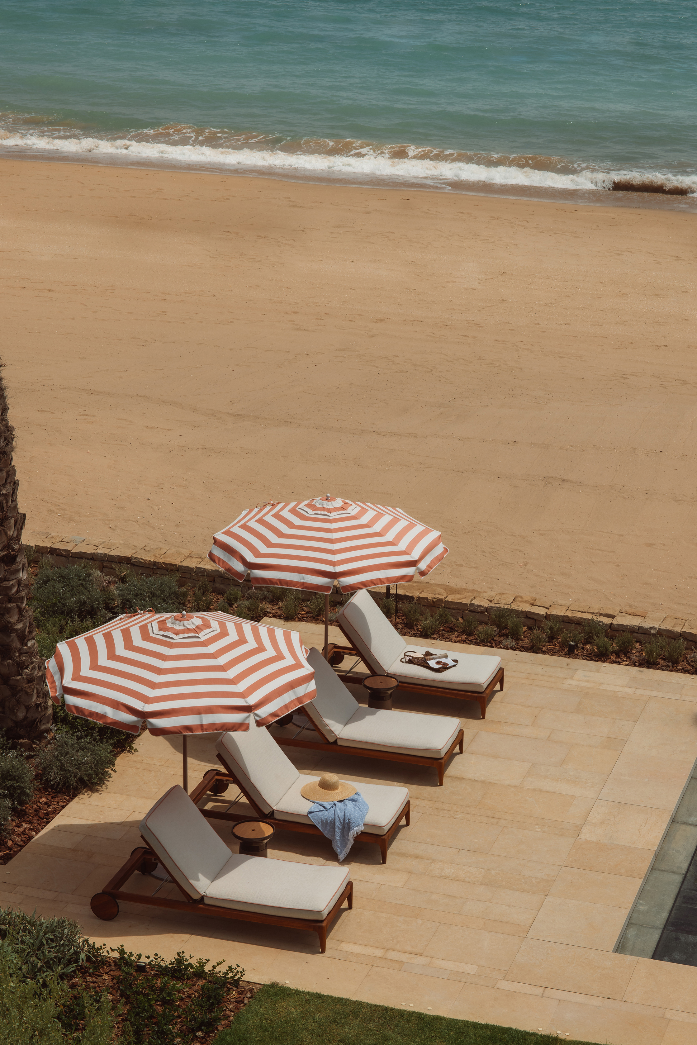 Four sun loungers with items scattered on them and shaded by red and white striped sun umbrellas, lined up in front of a beach