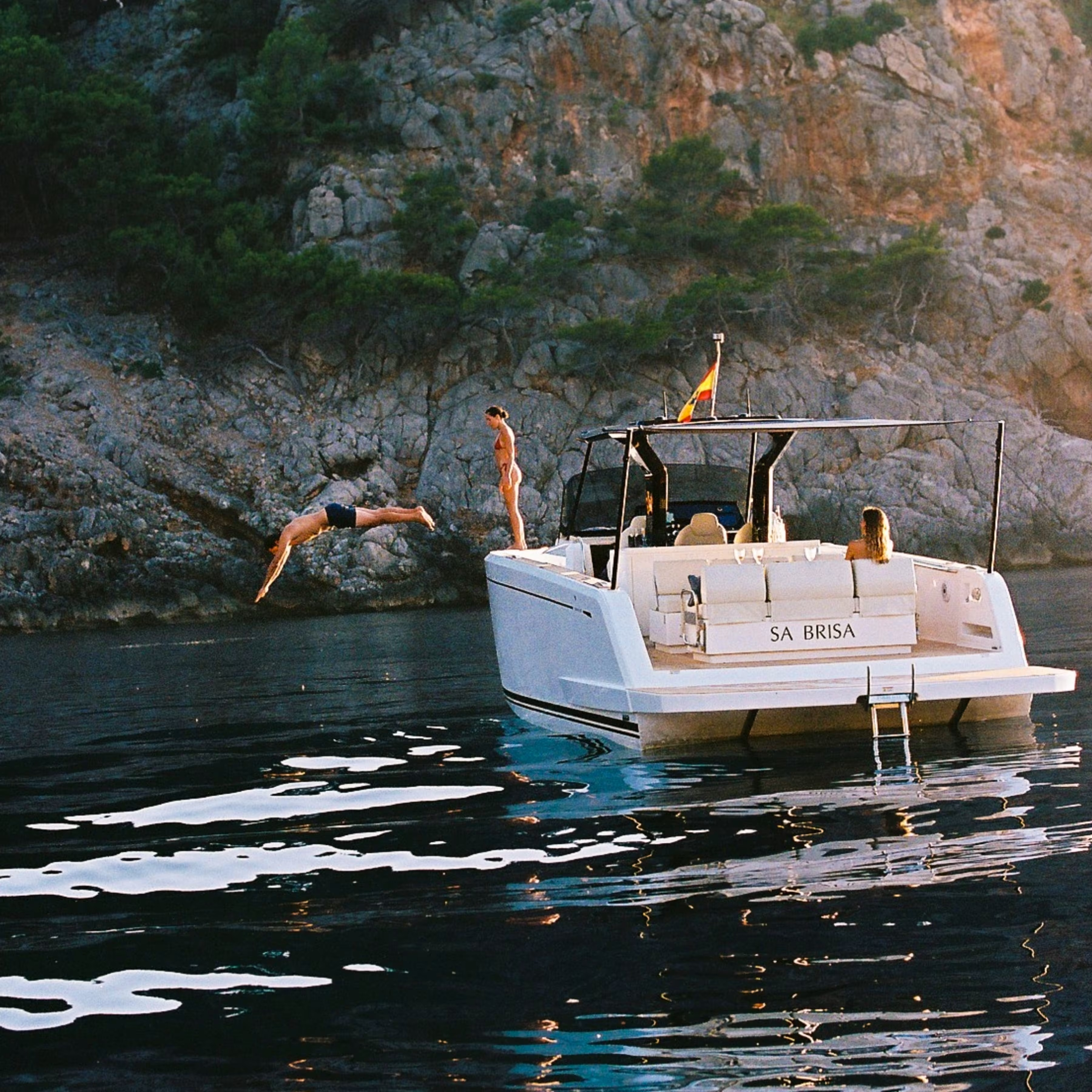 Luxury motorboat at La Residencia, a Belmond Hotel, anchored near rocky coastline with clear water, people enjoying swimming and diving."
