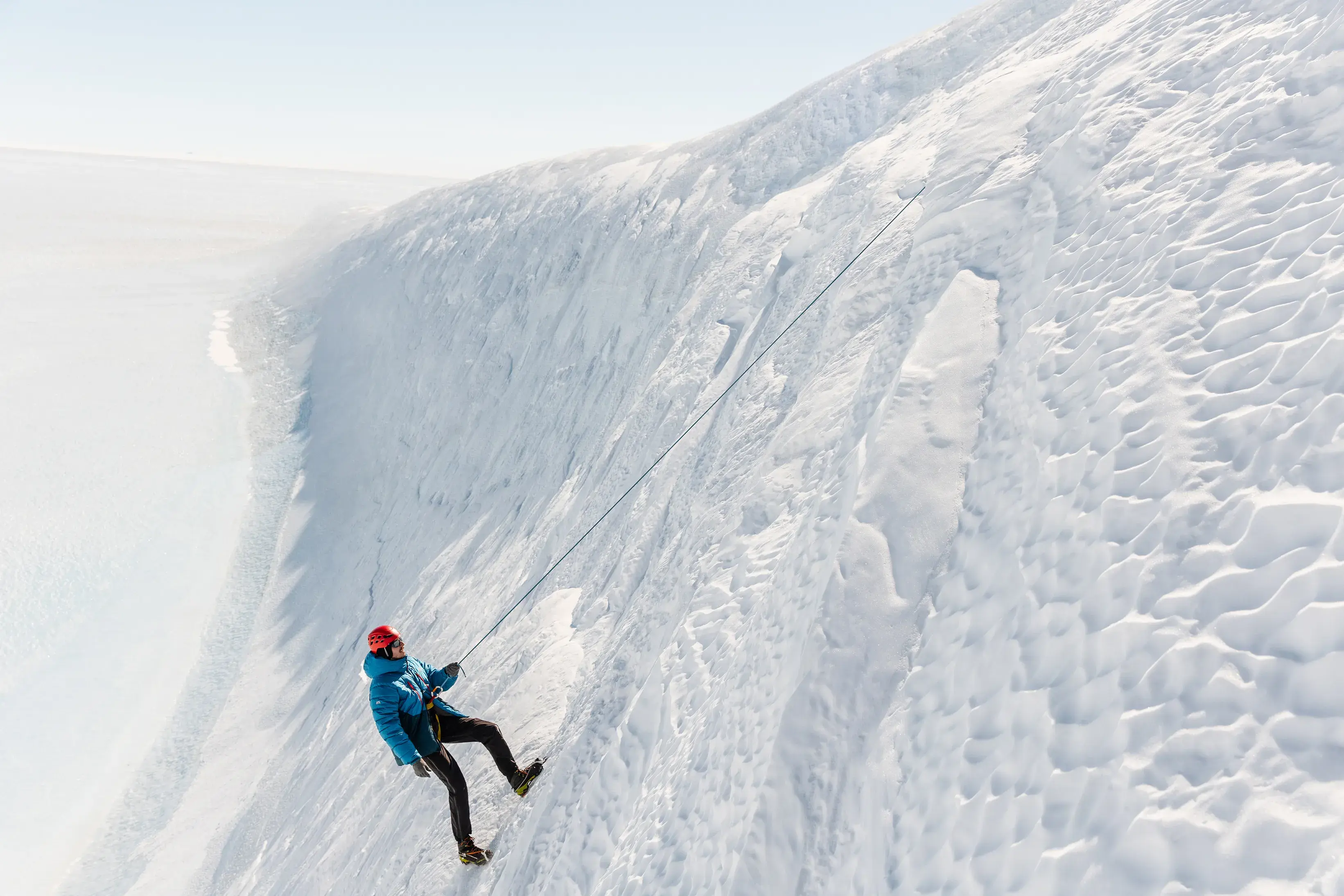 Person abseiling down a steep, textured ice wall in Antarctica under bright, clear conditions.