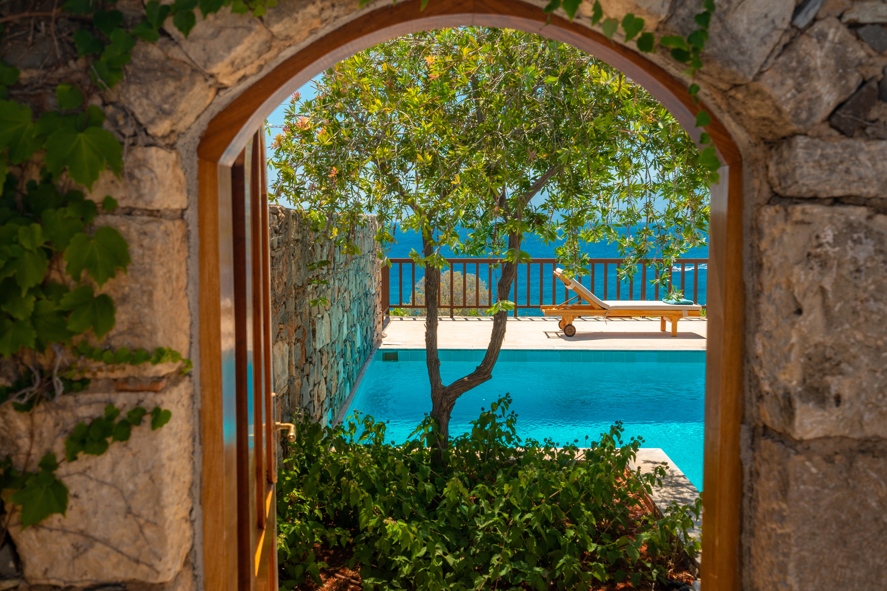 View through an archway to a pool with trees and sunloungers 