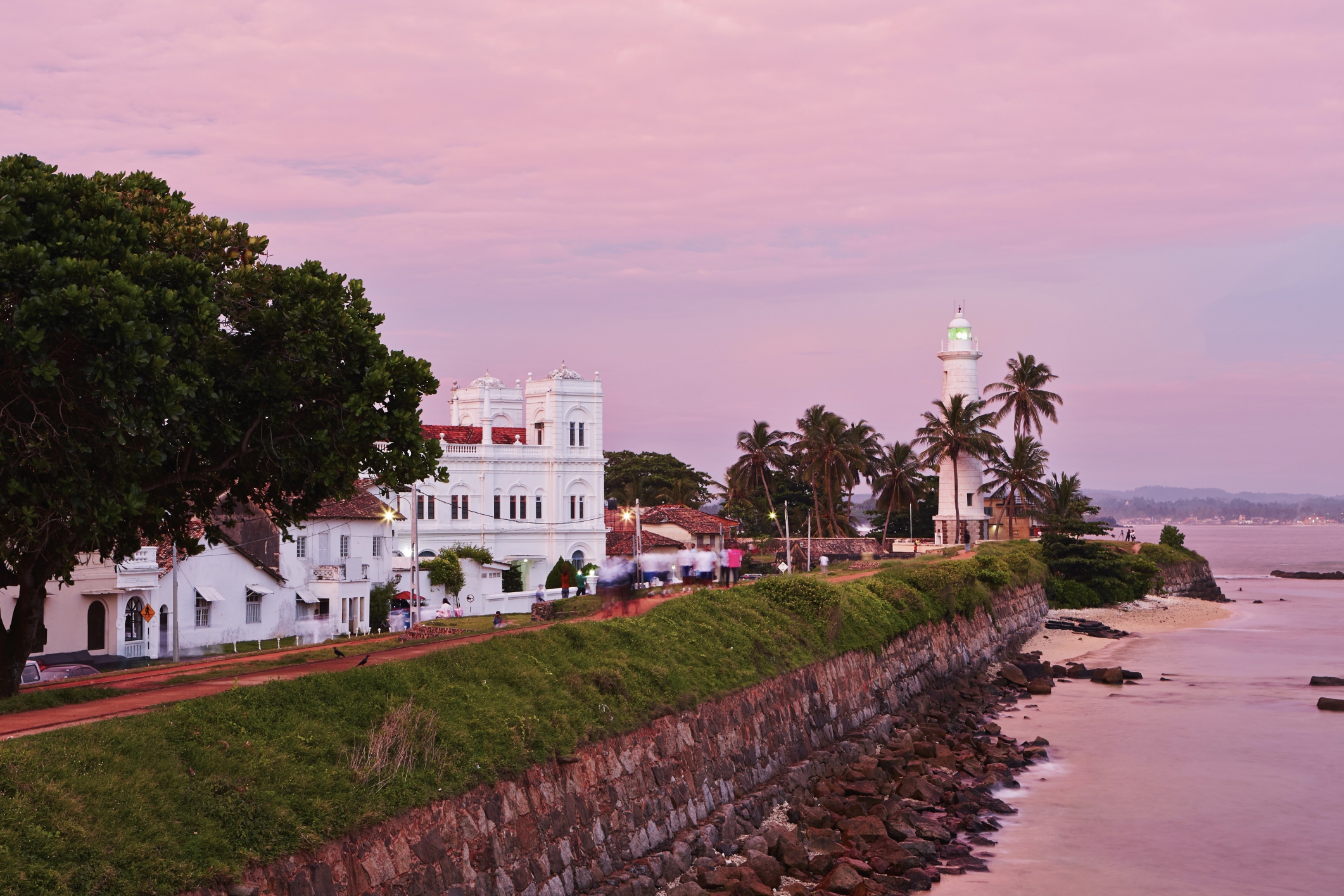 The Galle Fort walkway leading to a white lighthouse and colonial buildings at sunset with a pink sky over the ocean.