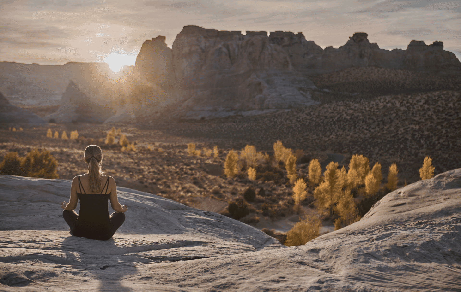 Person sitting cross‑legged in meditation on a rocky outcrop at sunrise, overlooking a desert canyon landscape.