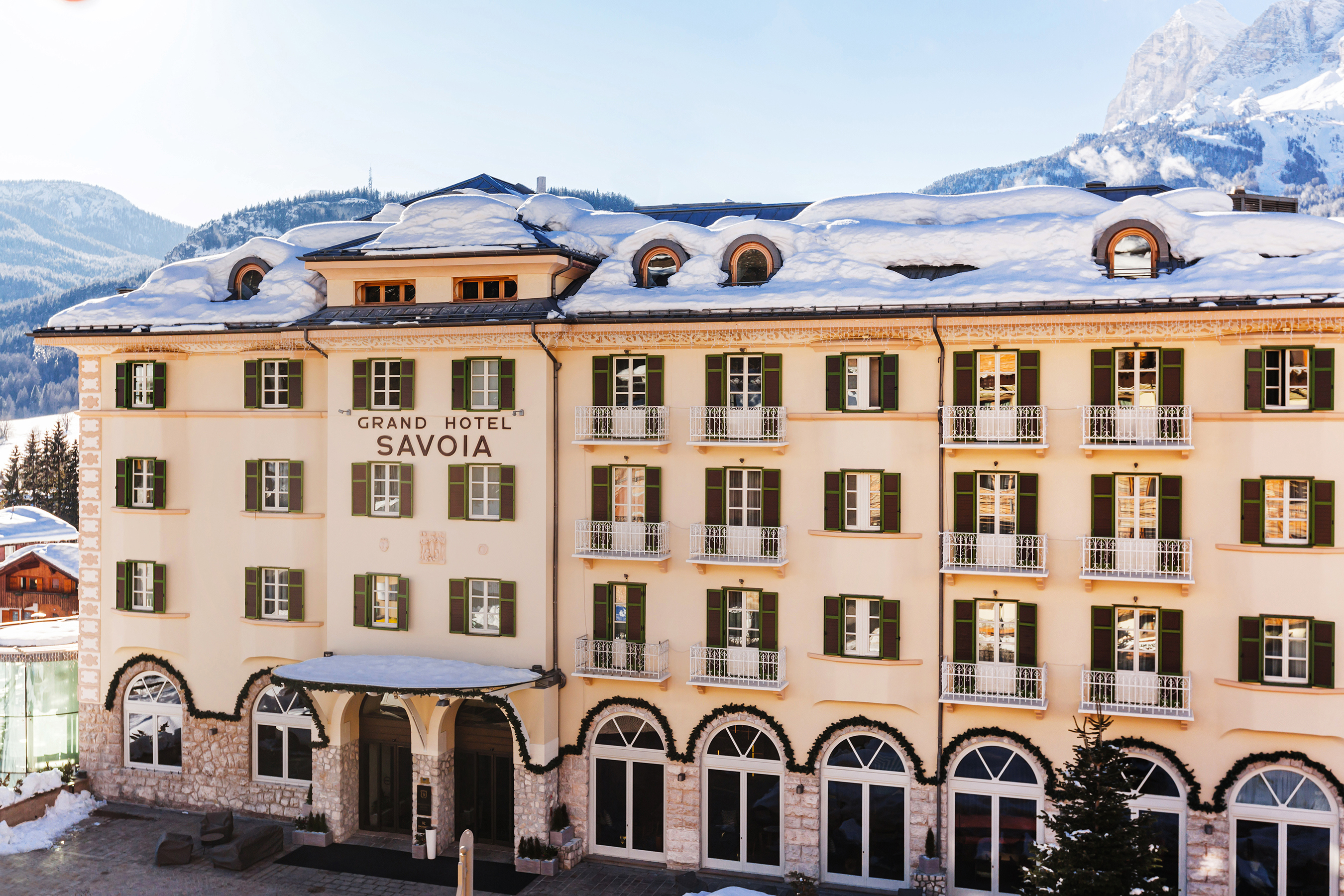The exterior view of Grand Hotel Savoia featuring a large cream building with lots of shuttered windows and snow on the roof backed by mountains under a clear sky