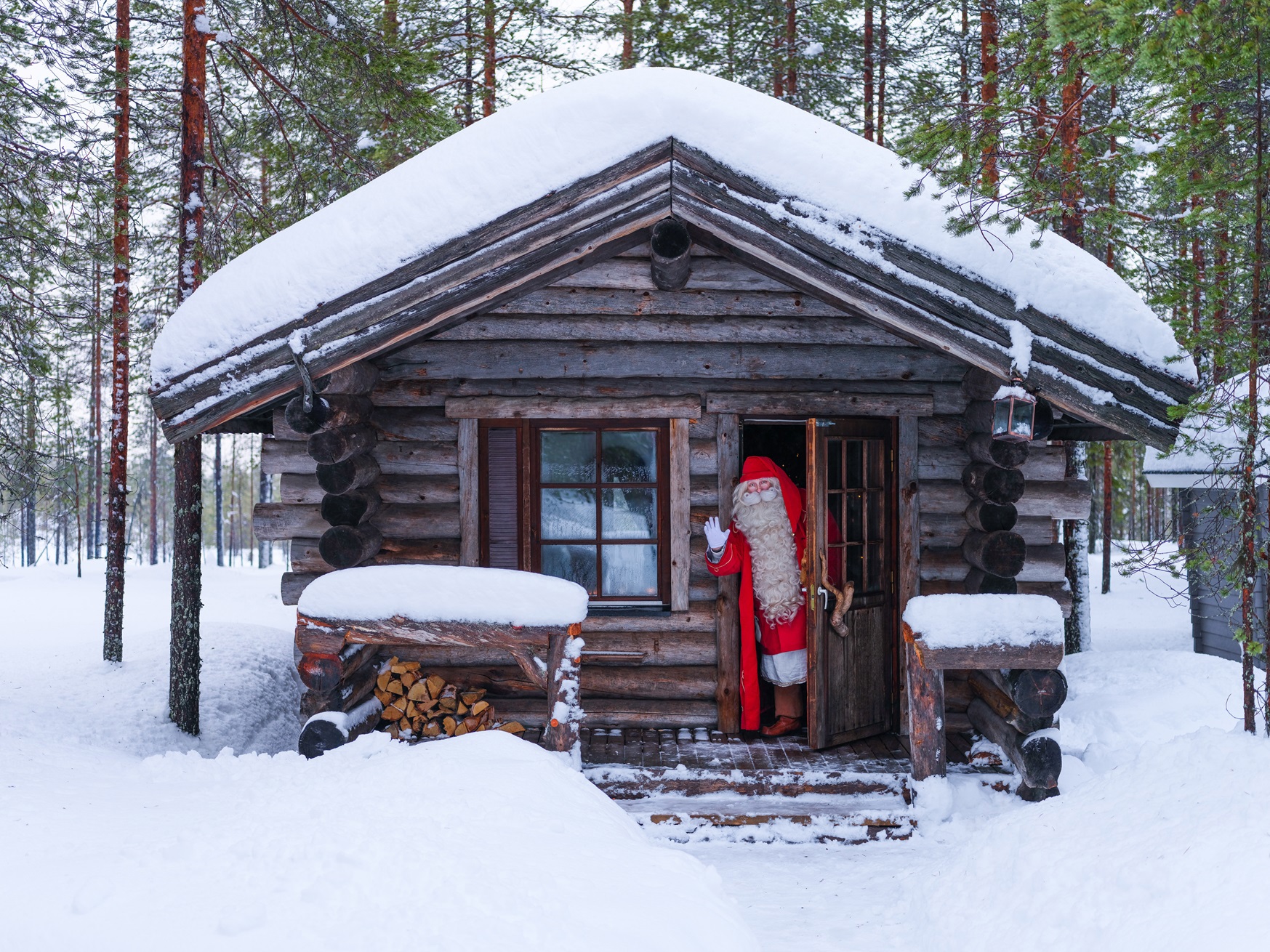 A person dressed as Santa Claus waving from the door of a log cabin covered in snow