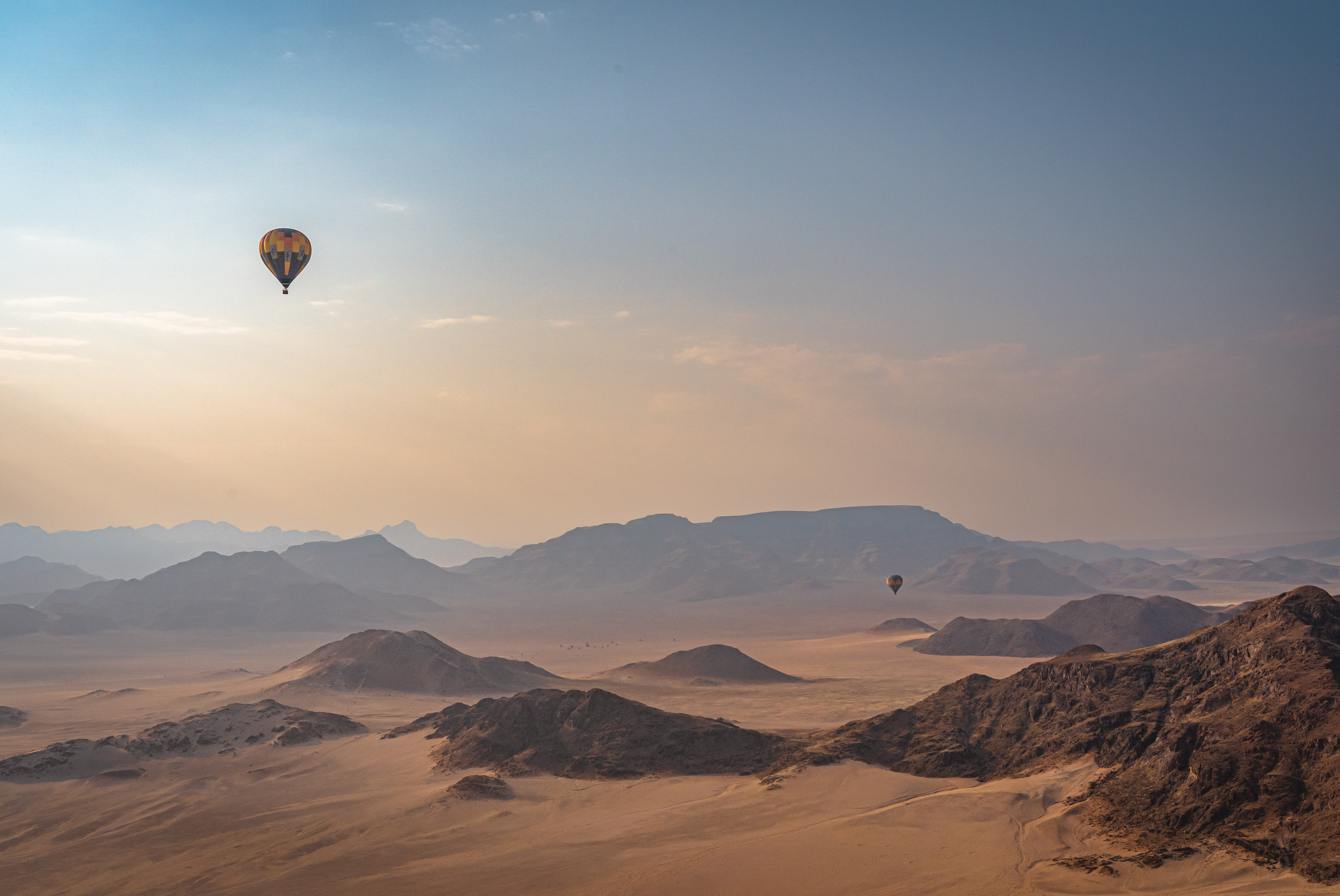 Two hot air balloons float over a vast Namibian desert landscape with mountains in the background under a clear sky