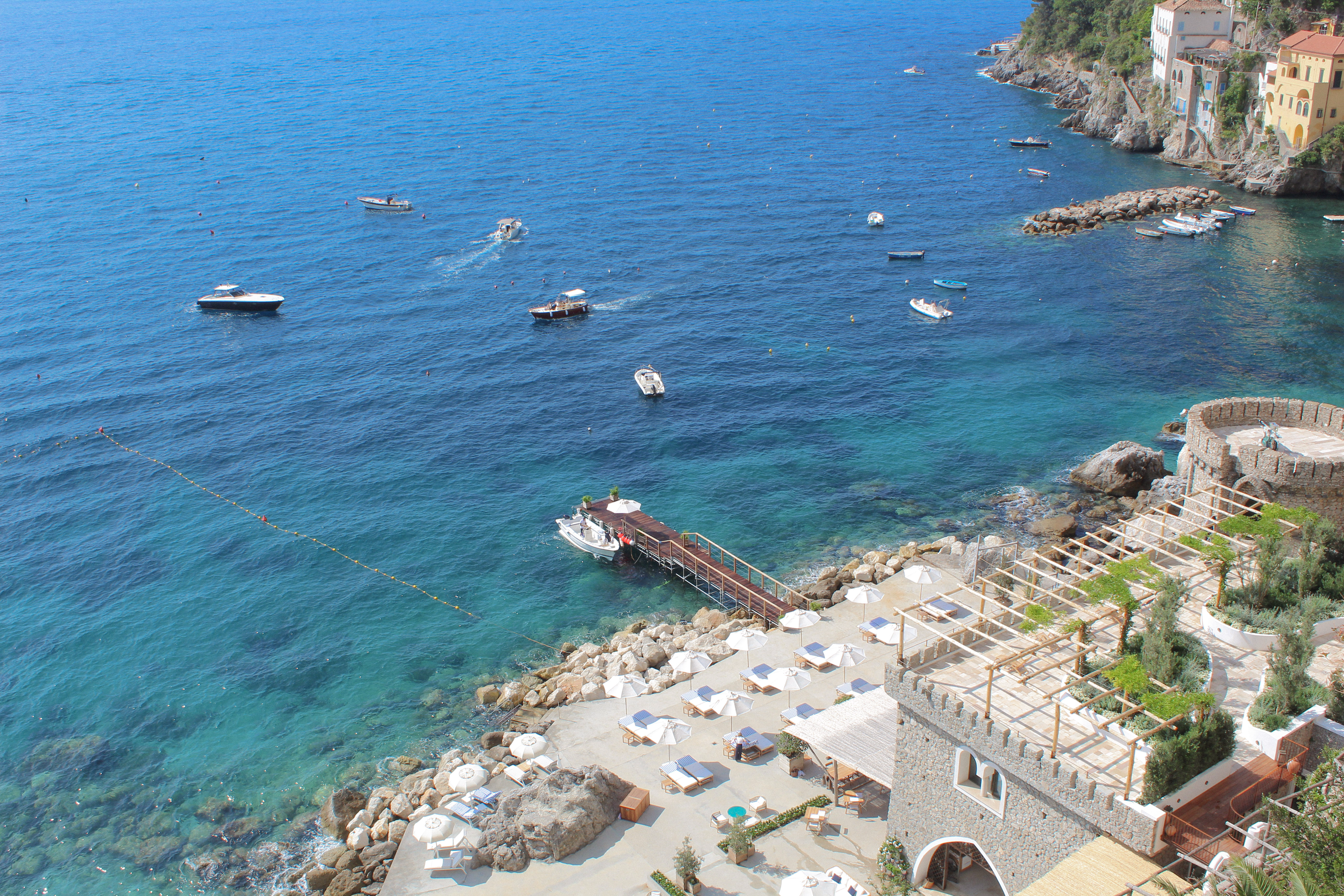 Aerial view of Borgo Santandrea's private beach and dock in Amalfi, showcasing turquoise waters, boats, and lush coastal landscape.
