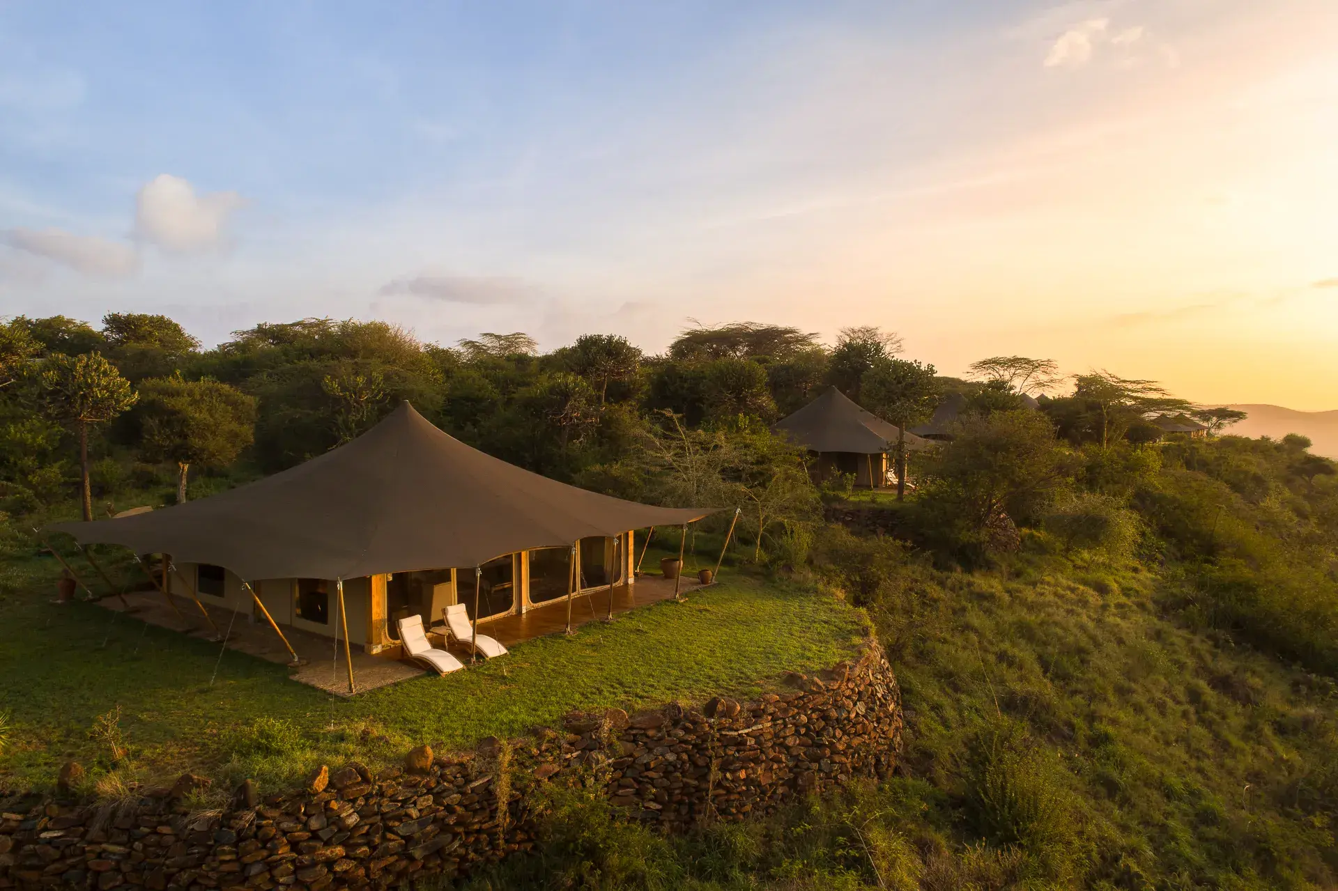 A luxury tent at Elewana Loisaba Tented Camp sitting on the edge of a cliff surrounded by trees with a glimpse of another tent behind at sunset