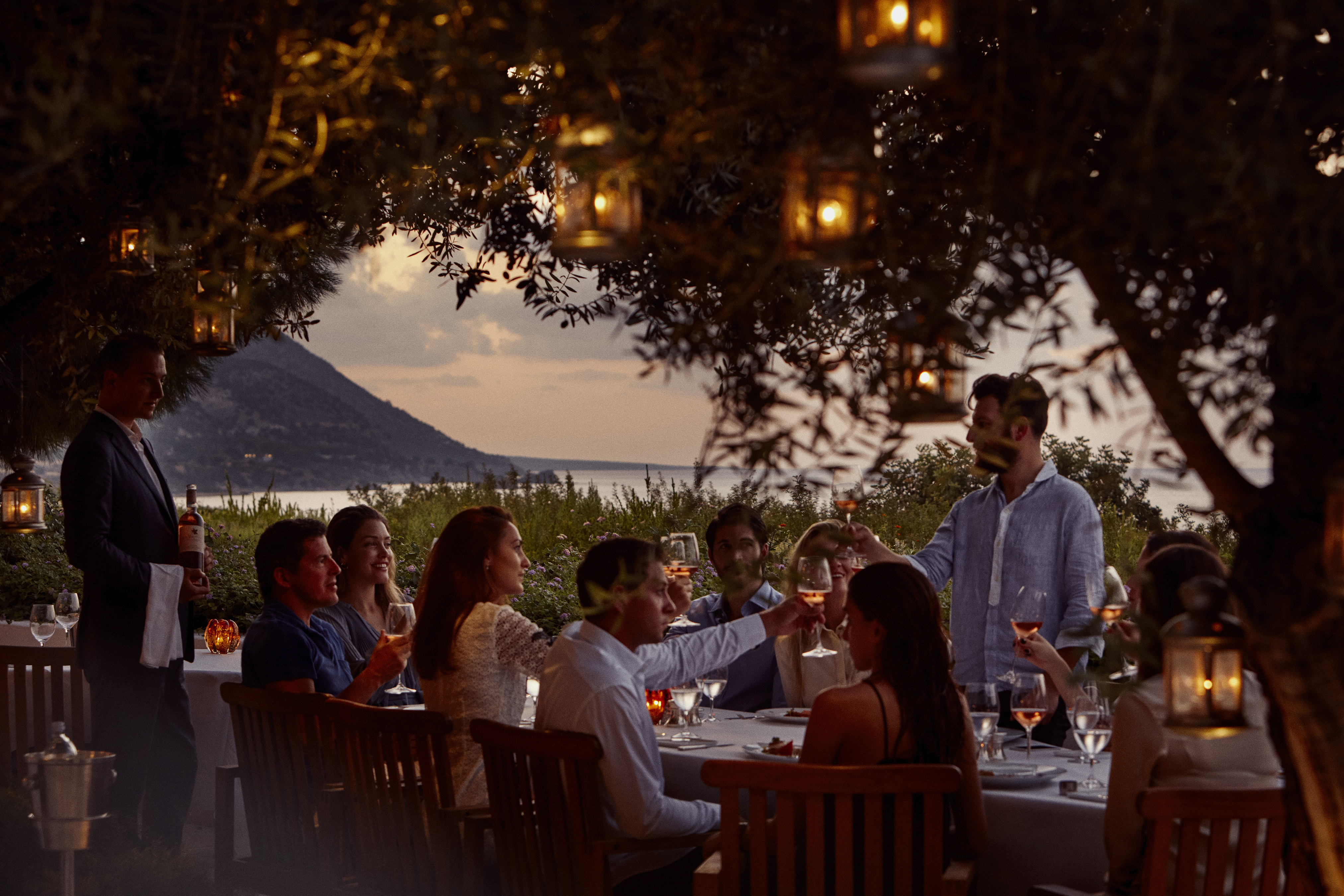 A group of guests dining outside at Helios Restaurant at Anassa in Cyprus on a table set beneath trees with lanterns and a view of the ocean and island behind
