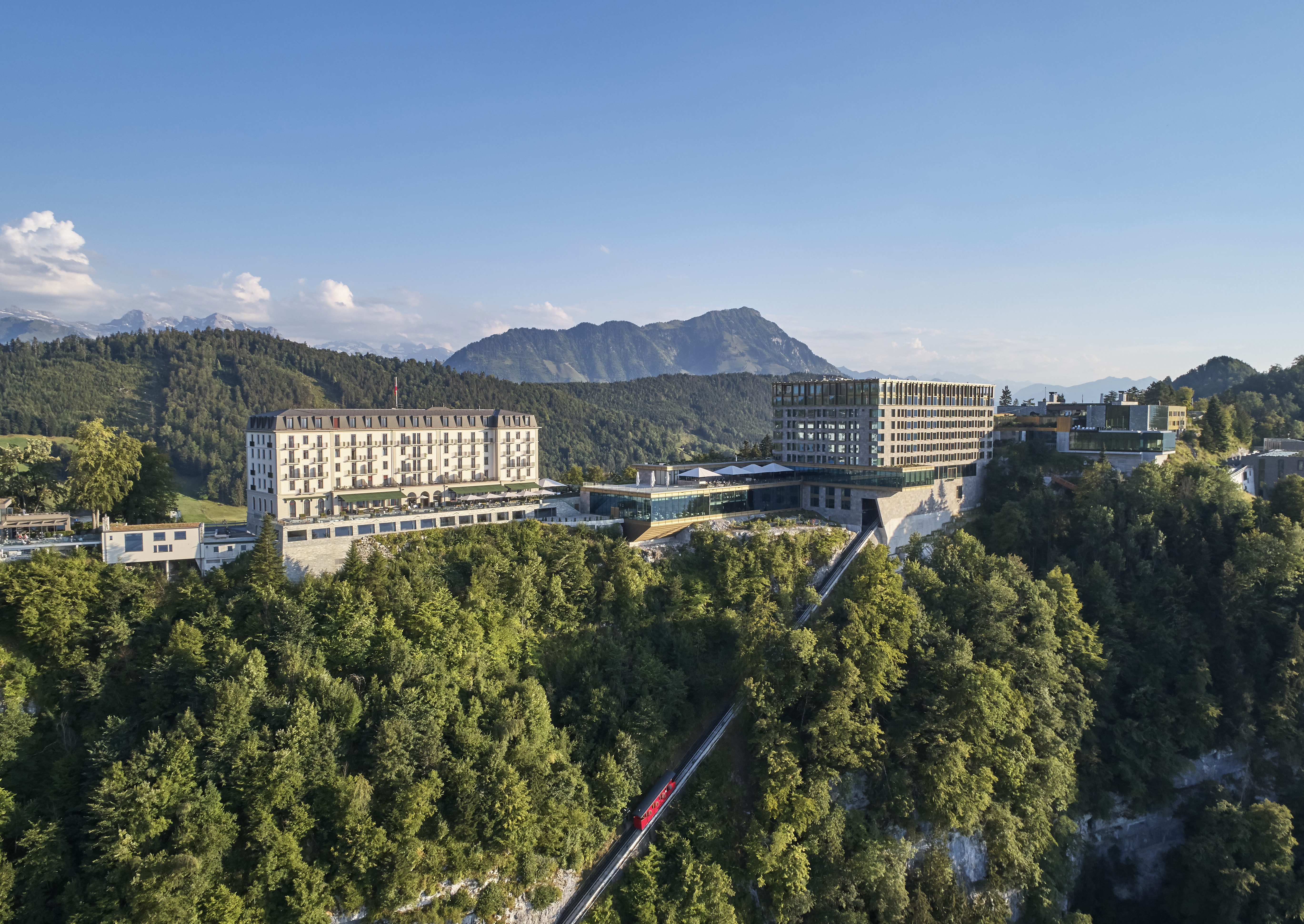 The modern exterior of Bürgenstock Hotel featuring two tiers of outdoor space and a red cable car arriving at the base of the hotel surrounded by greenery