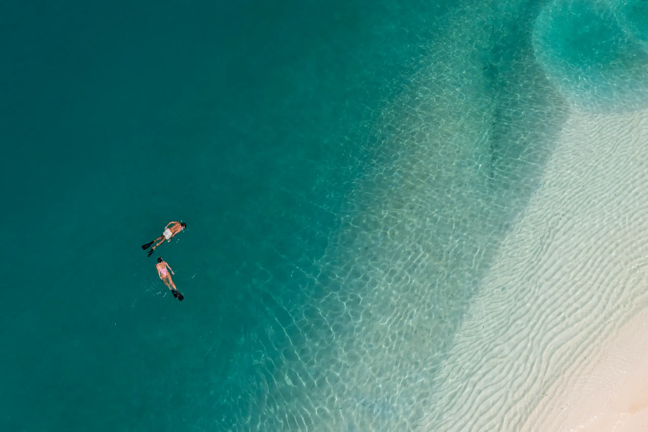 Two people snorkeling in clear turquoise water near a sandy beach.