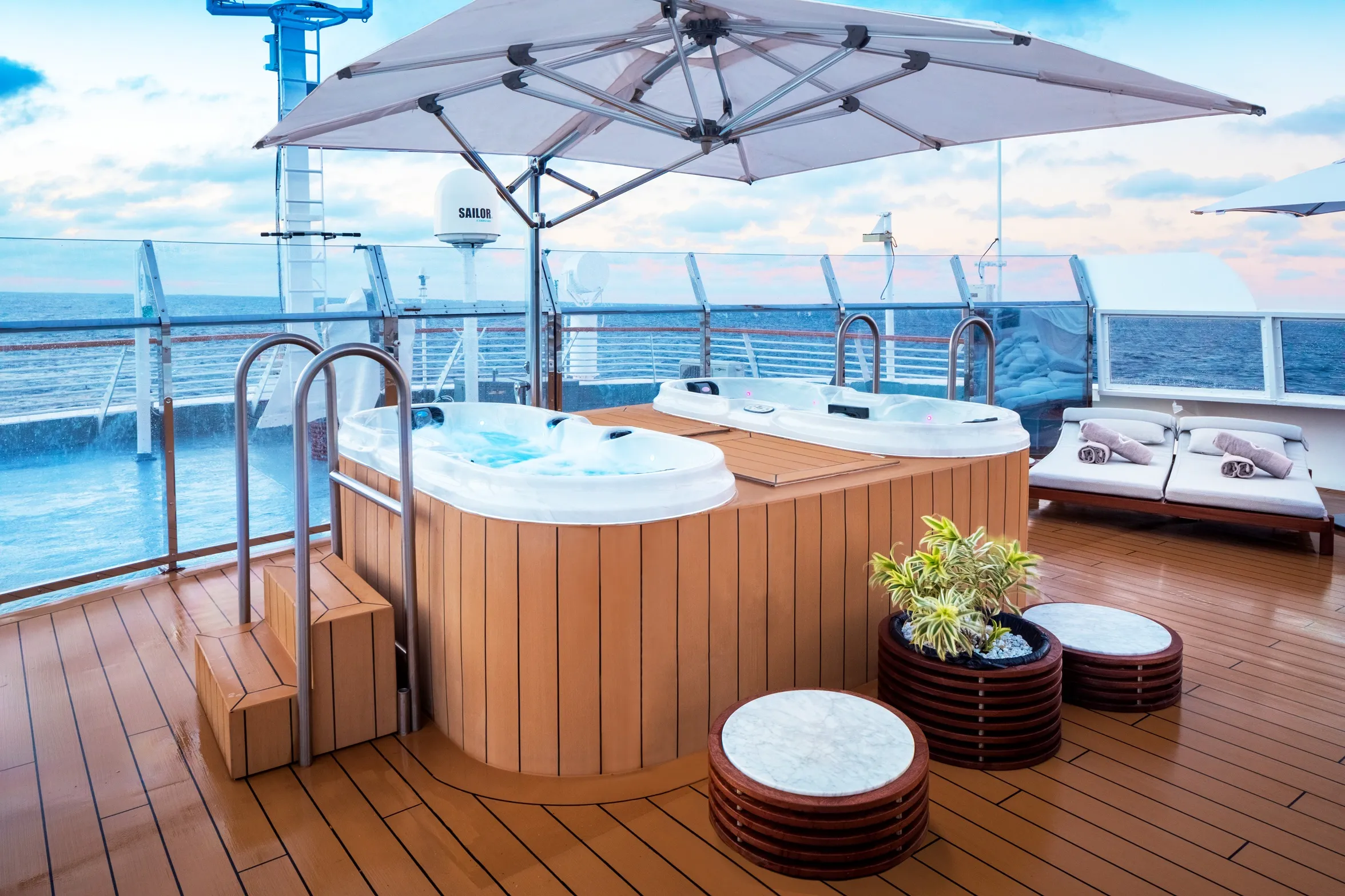 Cruise ship deck with two hot tubs, lounge chairs, and ocean views under a large umbrella.