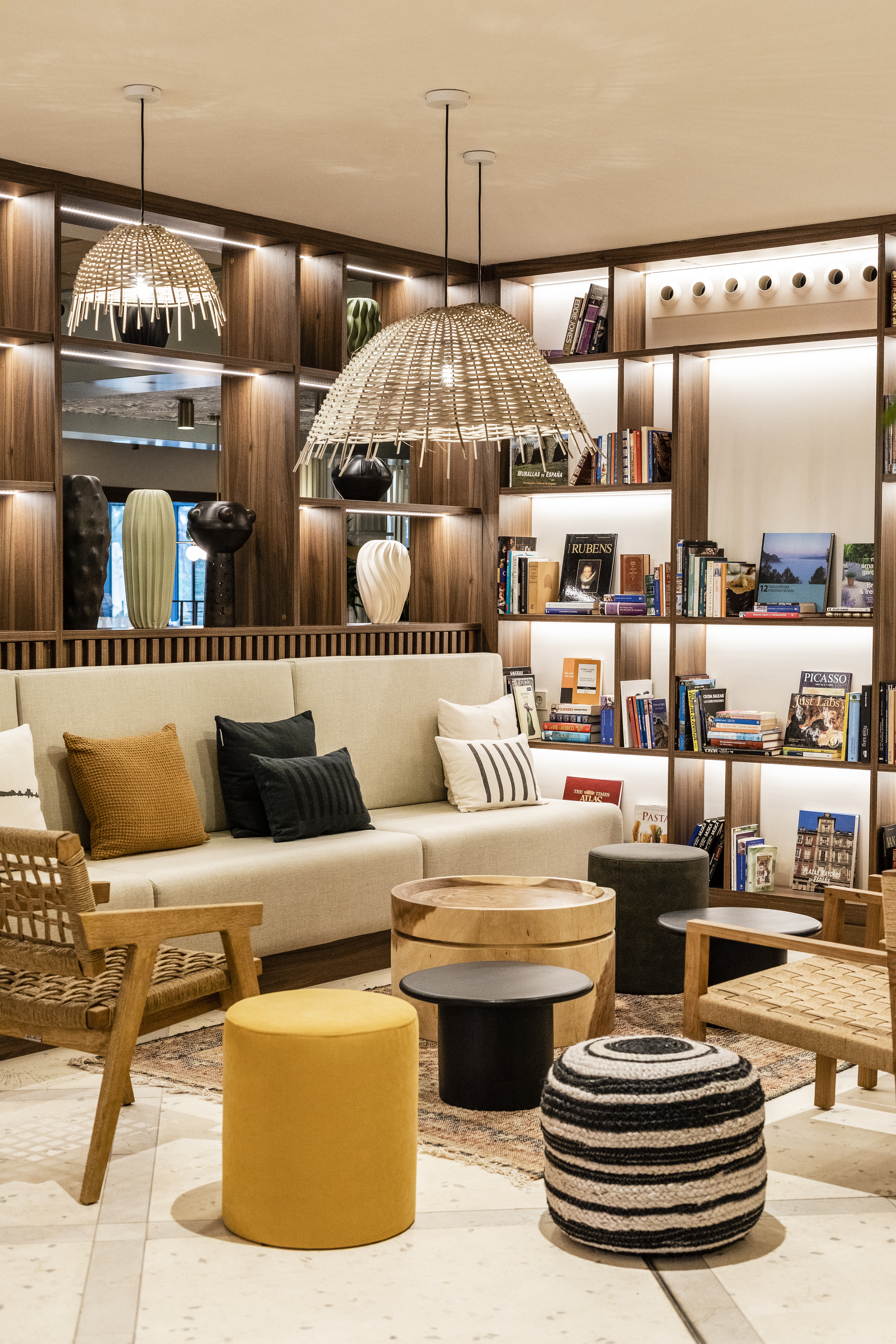 A cozy living room with a beige sofa, wooden bookshelves filled with books, a wicker armchair, and various decorative elements including a yellow ottoman and striped stool under warm lighting