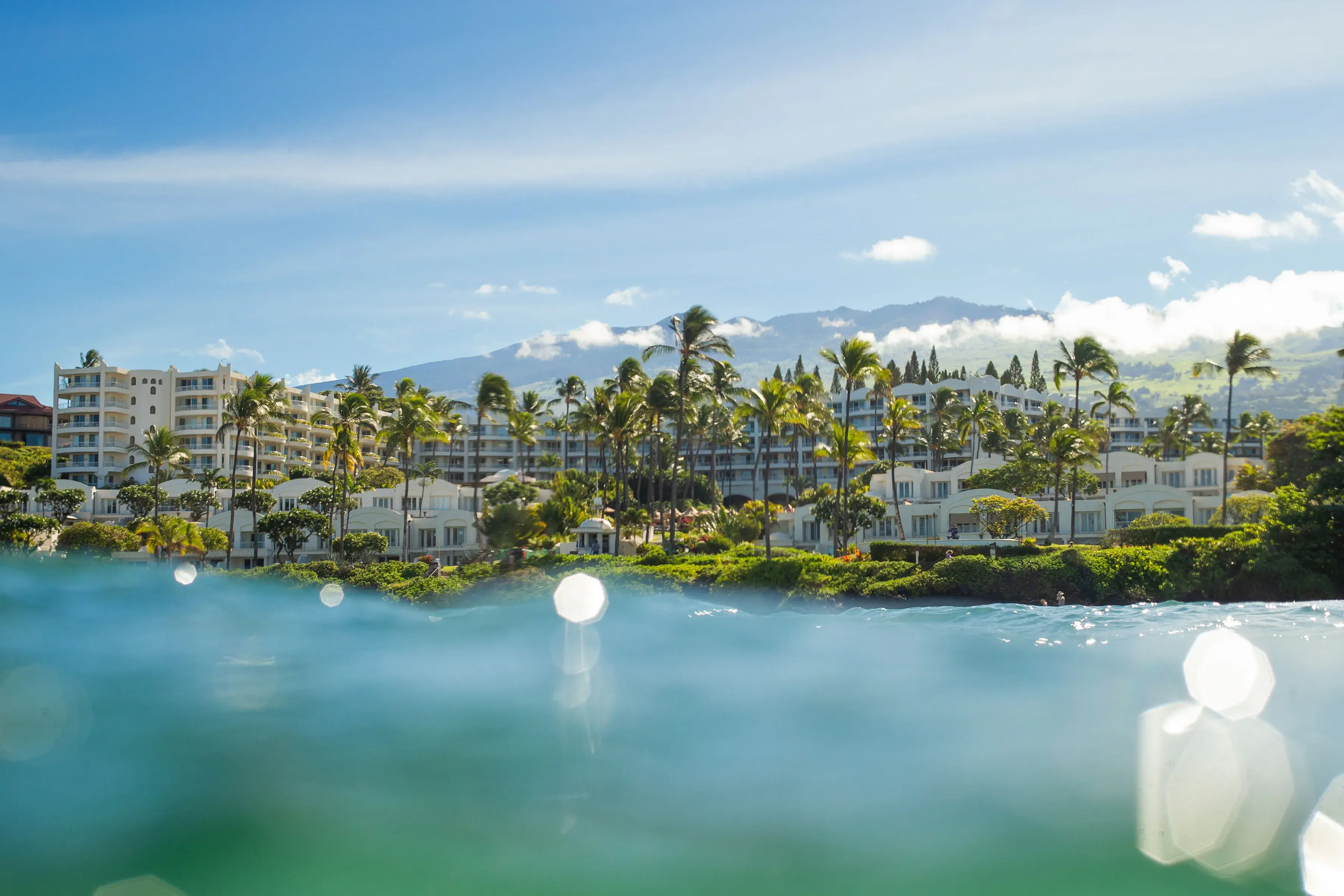 The view of Fairmont Kea Lani from the water featuring the hotel surrounded by palms and backed by mountains, with the surface of the water blurred in the foreground