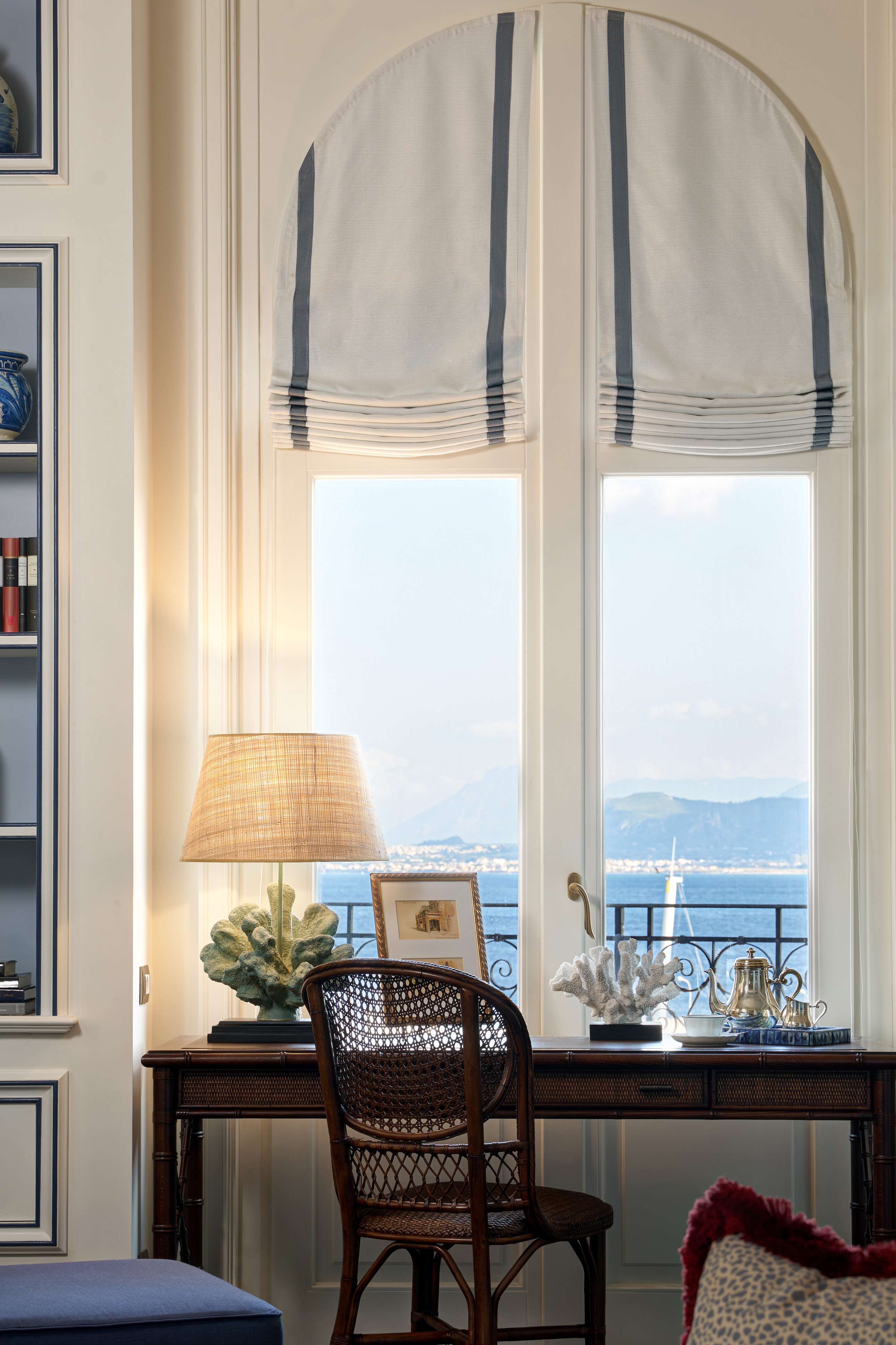 A well-lit window area with classic decor featuring a wooden desk, a woven chair, and a lamp, with the open window overlooking a scenic mountain view.