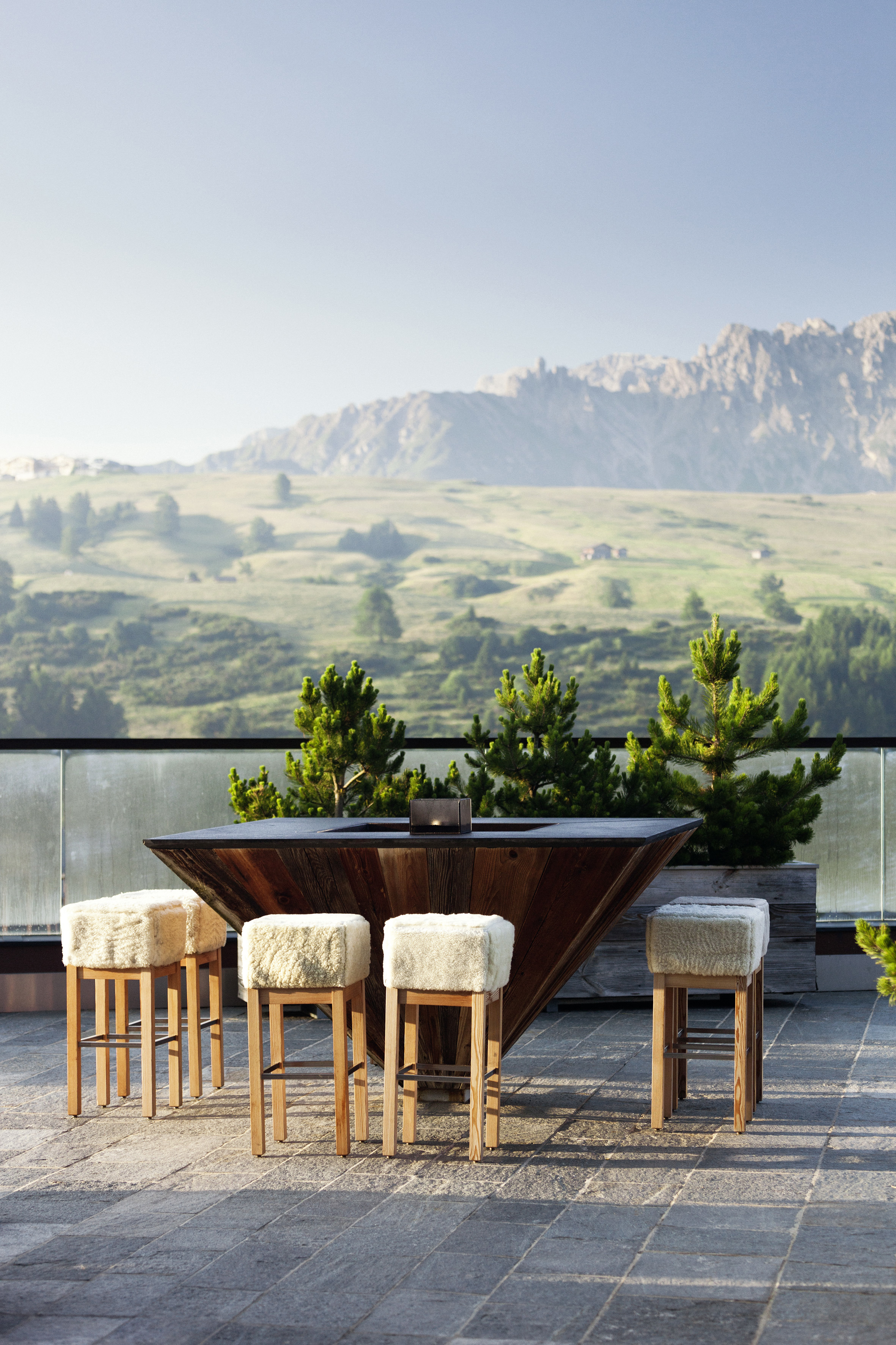 COMO Alpina outdoor dining area with a wooden table and fluffy stools overlooking a scenic hilly landscape under a clear blue summer sky.