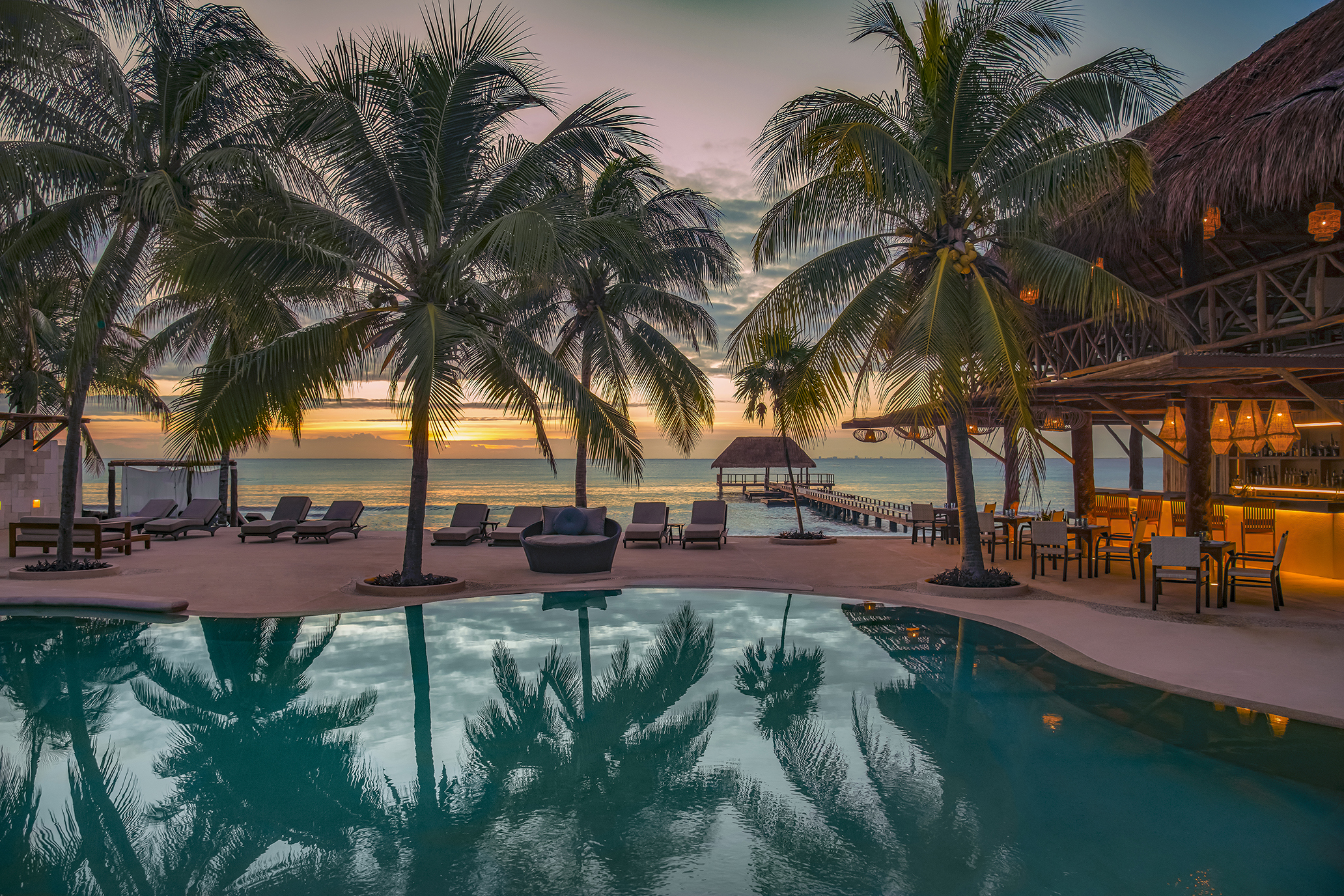 A pool flanked by palms and loungers and a thatched roof bar lit up at sunset
