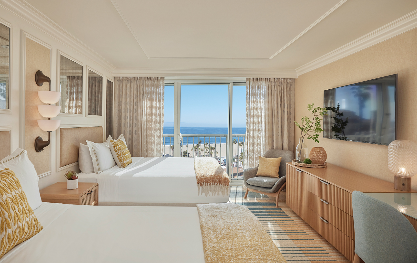 A twin hotel suite with a window view of a blue sky and ocean. The room is decorated in cream and brown tones and features twin double beds, a large chest of drawers, a TV and two blue arm chairs.