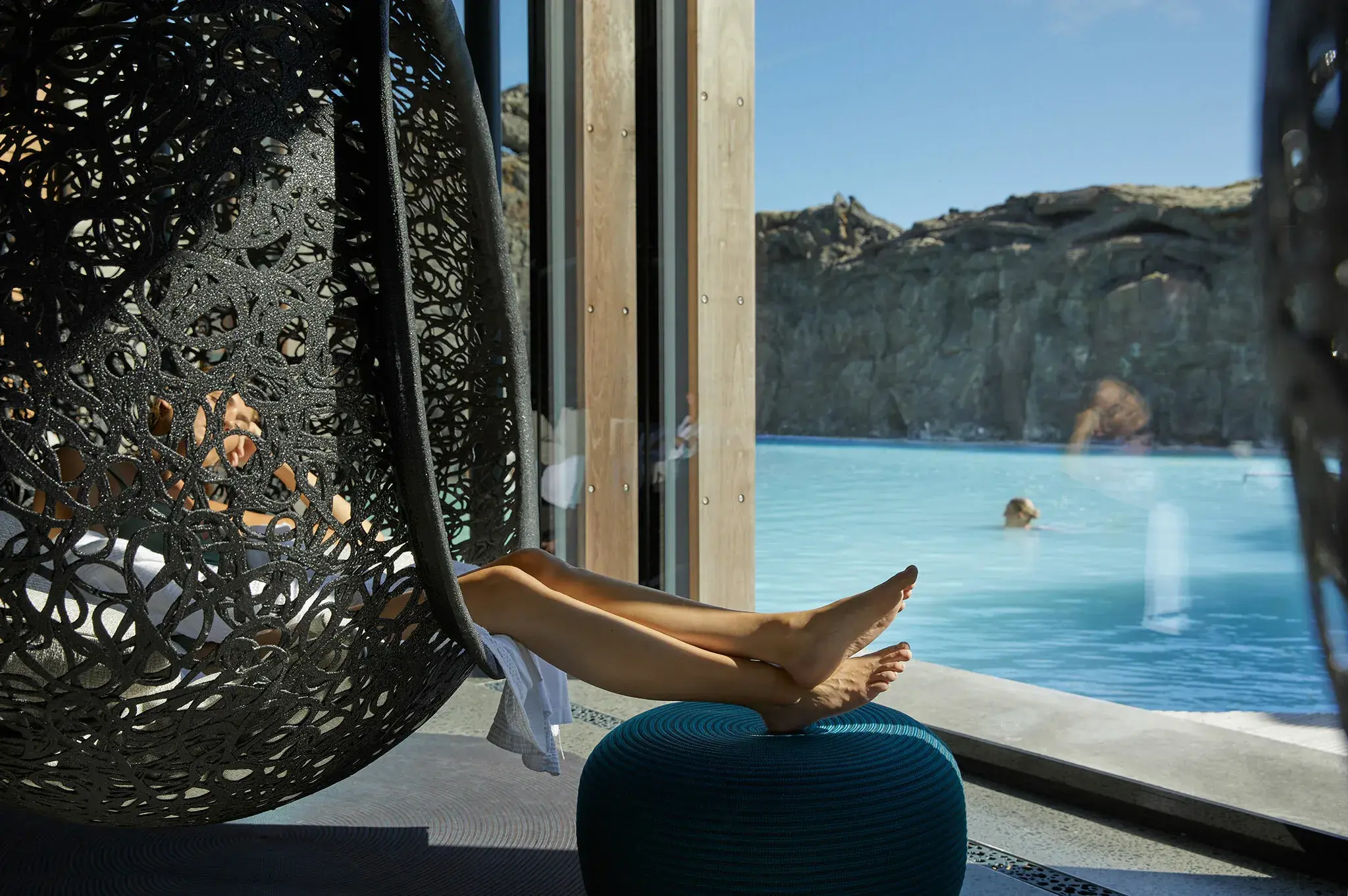A woman lounging in a hanging chair with only her legs visible in front of a large window with a woman swimming in the lagoon outside