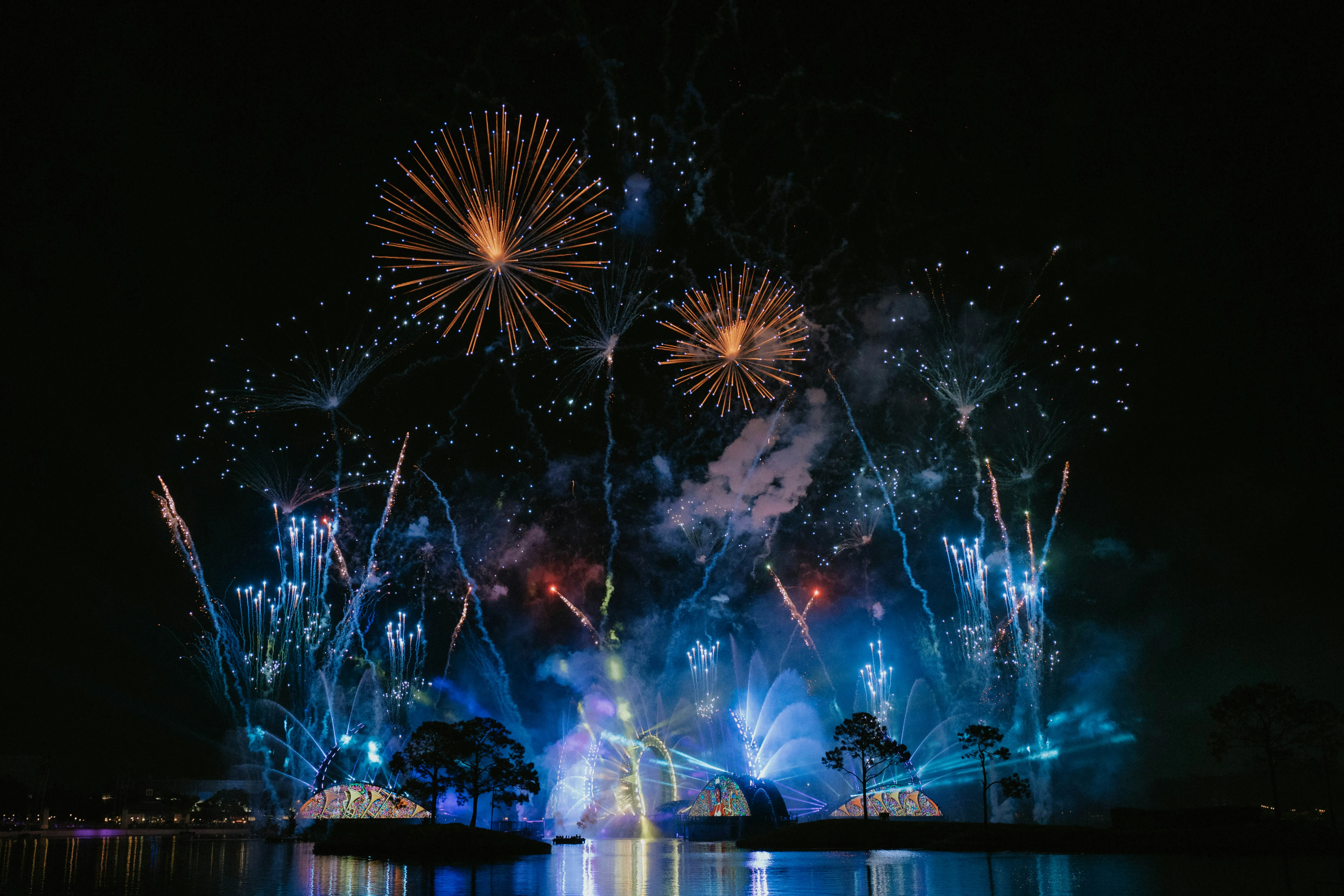 Colourful fireworks bursting over a nighttime waterfront with illuminated structures reflected in the water.