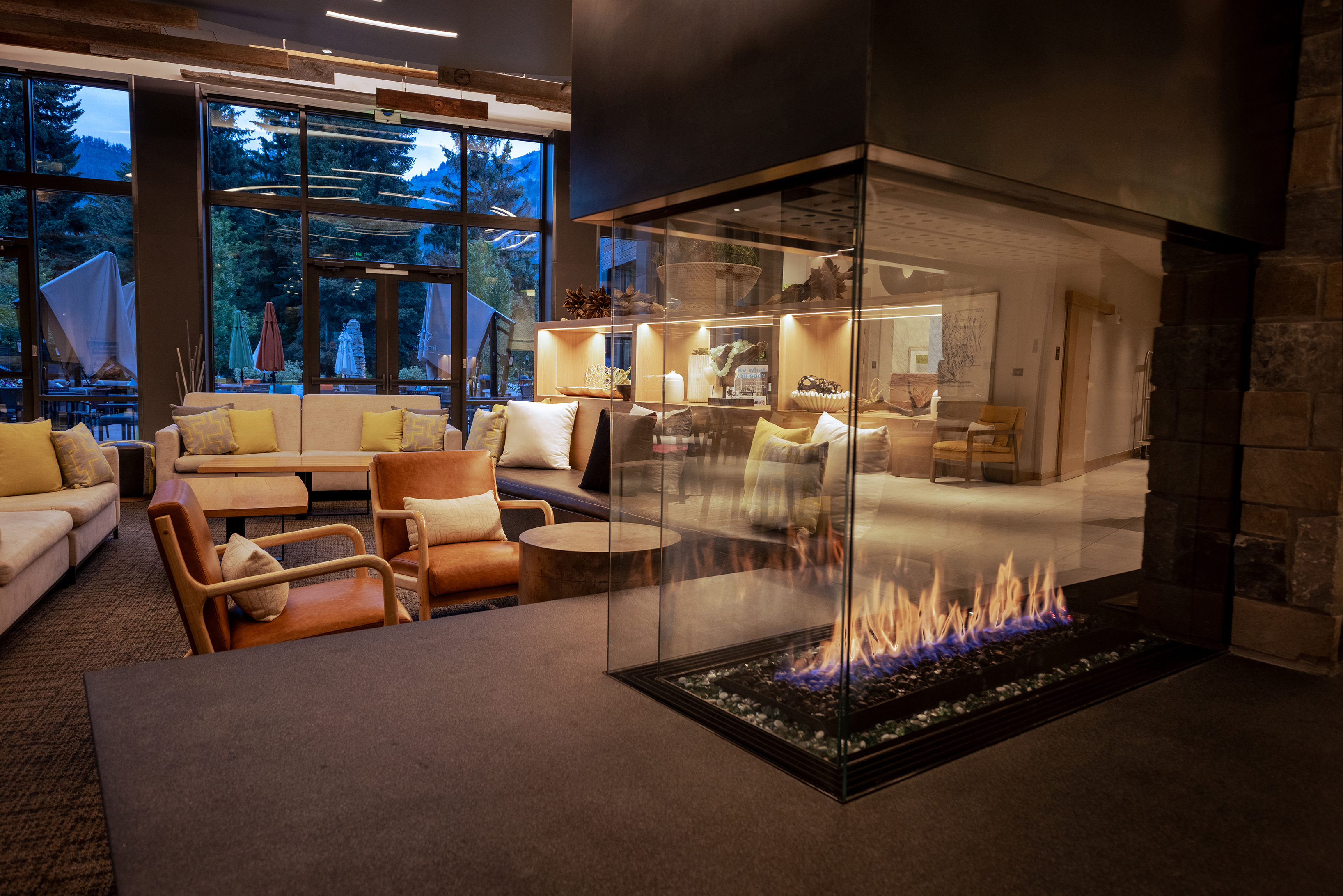 A cosy lounge area at Limelight Ketchum hotel featuring modern furniture, a central glass-encased fireplace, and large windows with an evening outdoor view
