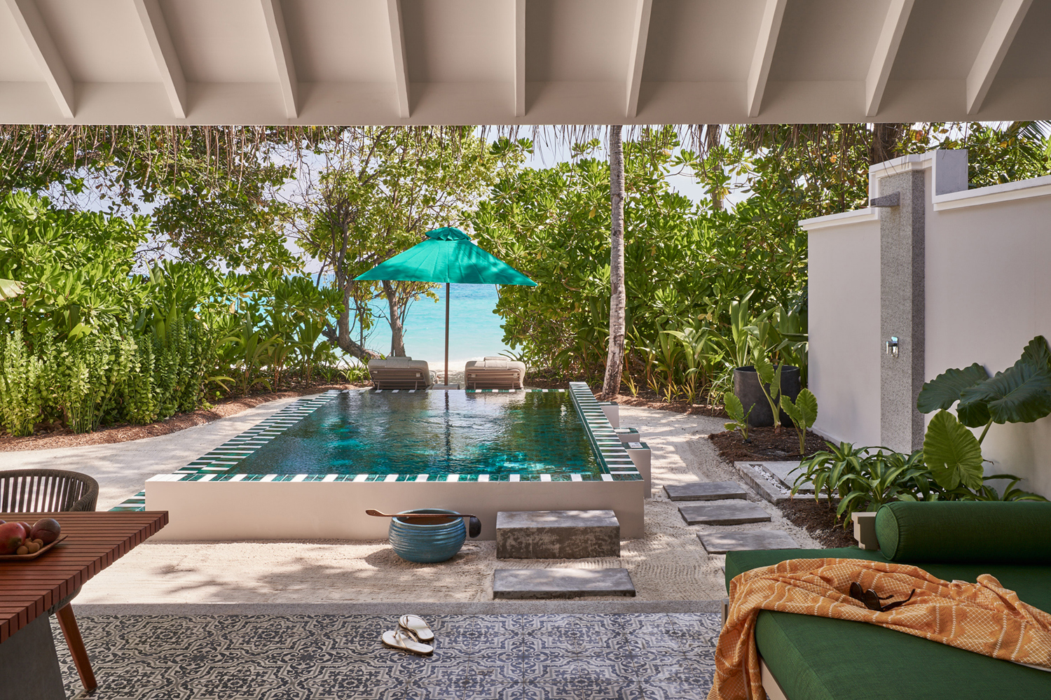 A view from the terrace of a Beach Pool Villa featuring a glimpse of a sofa and table before a private plunge pool with sun loungers and the beach and greenery beyond