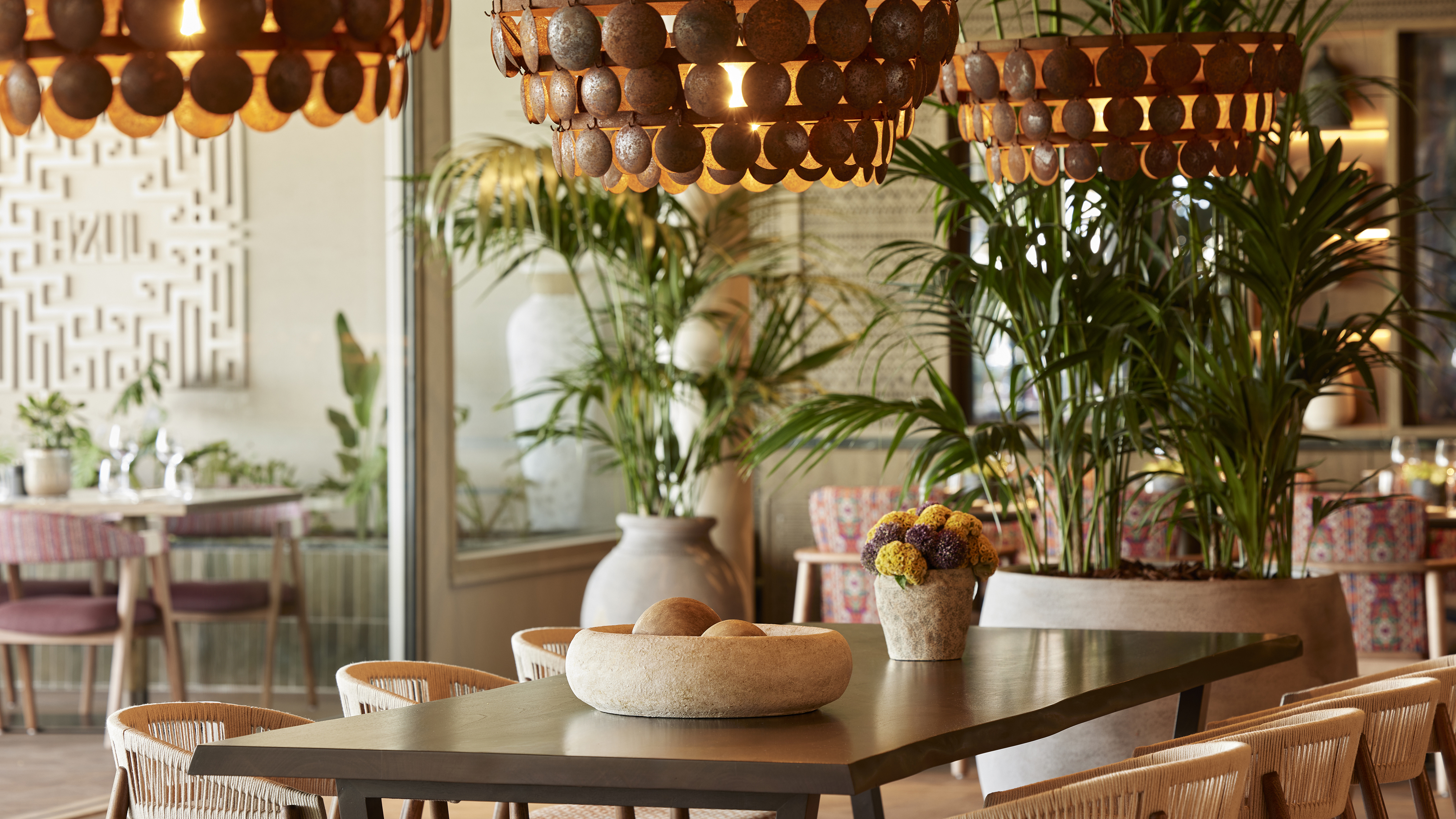 The interior of Azul Restaurant at Ikos Odissia featuring a dark wood table with decor beneath beaded lights and large potted plants in the background