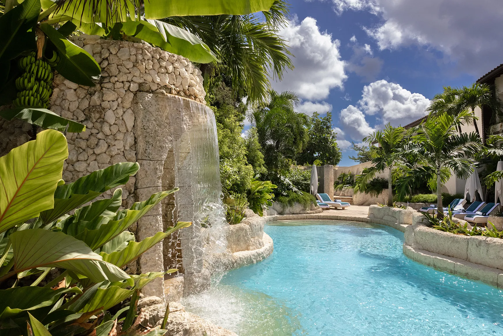 Tropical resort pool with a waterfall feature, lush greenery, and sun loungers surrounded by palm trees.