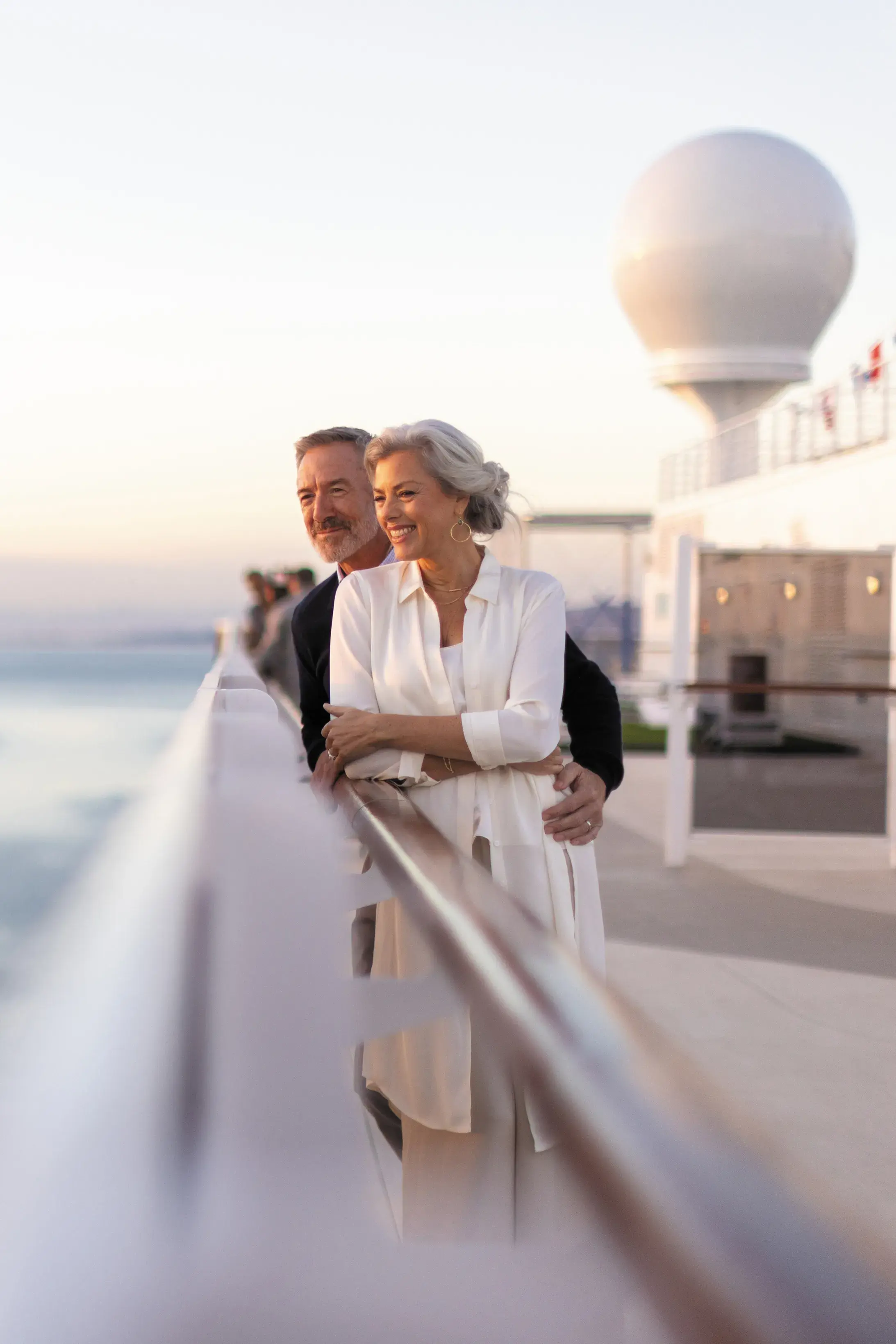 Couple standing on a Regent Seven Seas cruise ship deck overlooking the ocean at sunset with modern radar dome in the background.