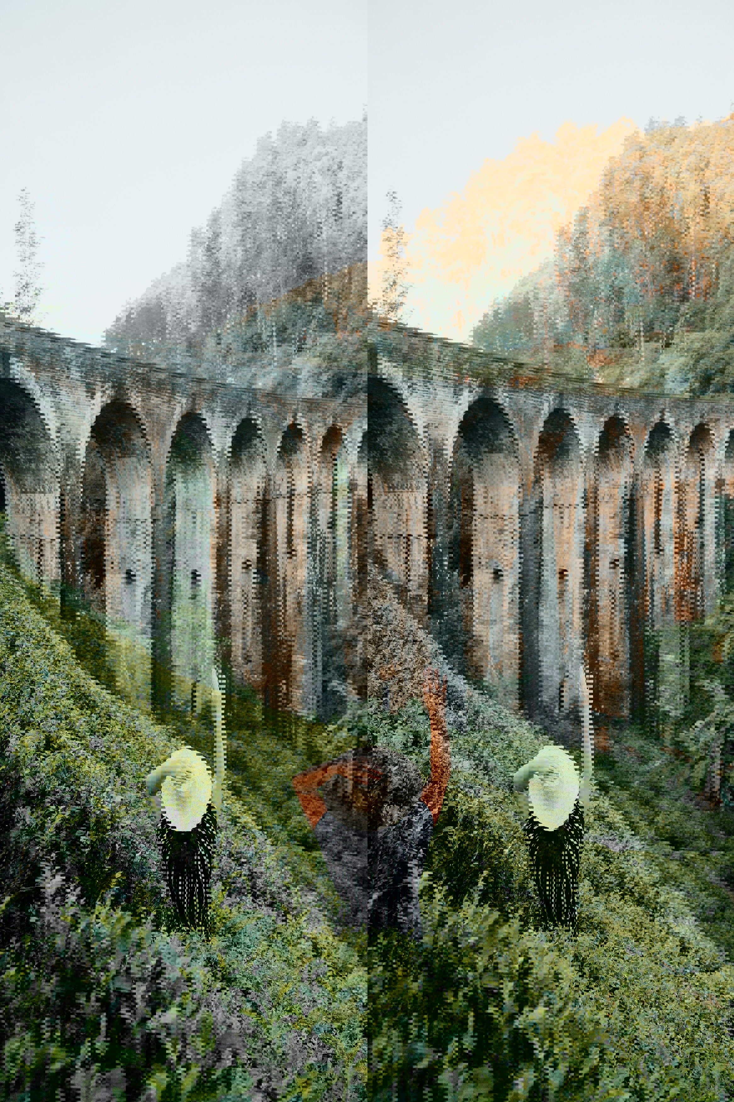 Woman in striped shirt and white sun hat standing amongst greenery holding up her hand towards an arhced railway bridge