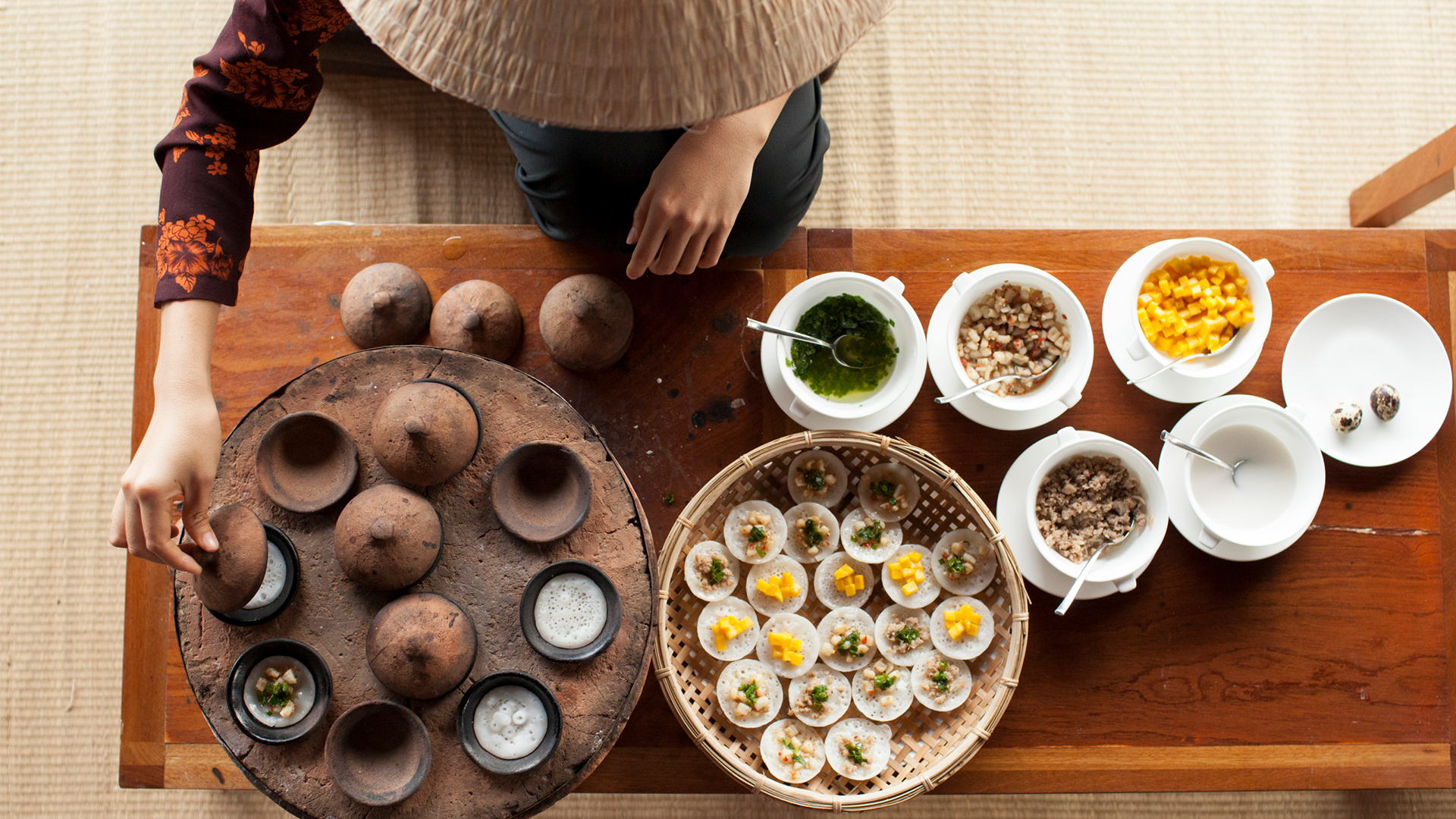 A birdseye view of table full of bowls of Asian food served by a women wearing a conical hat