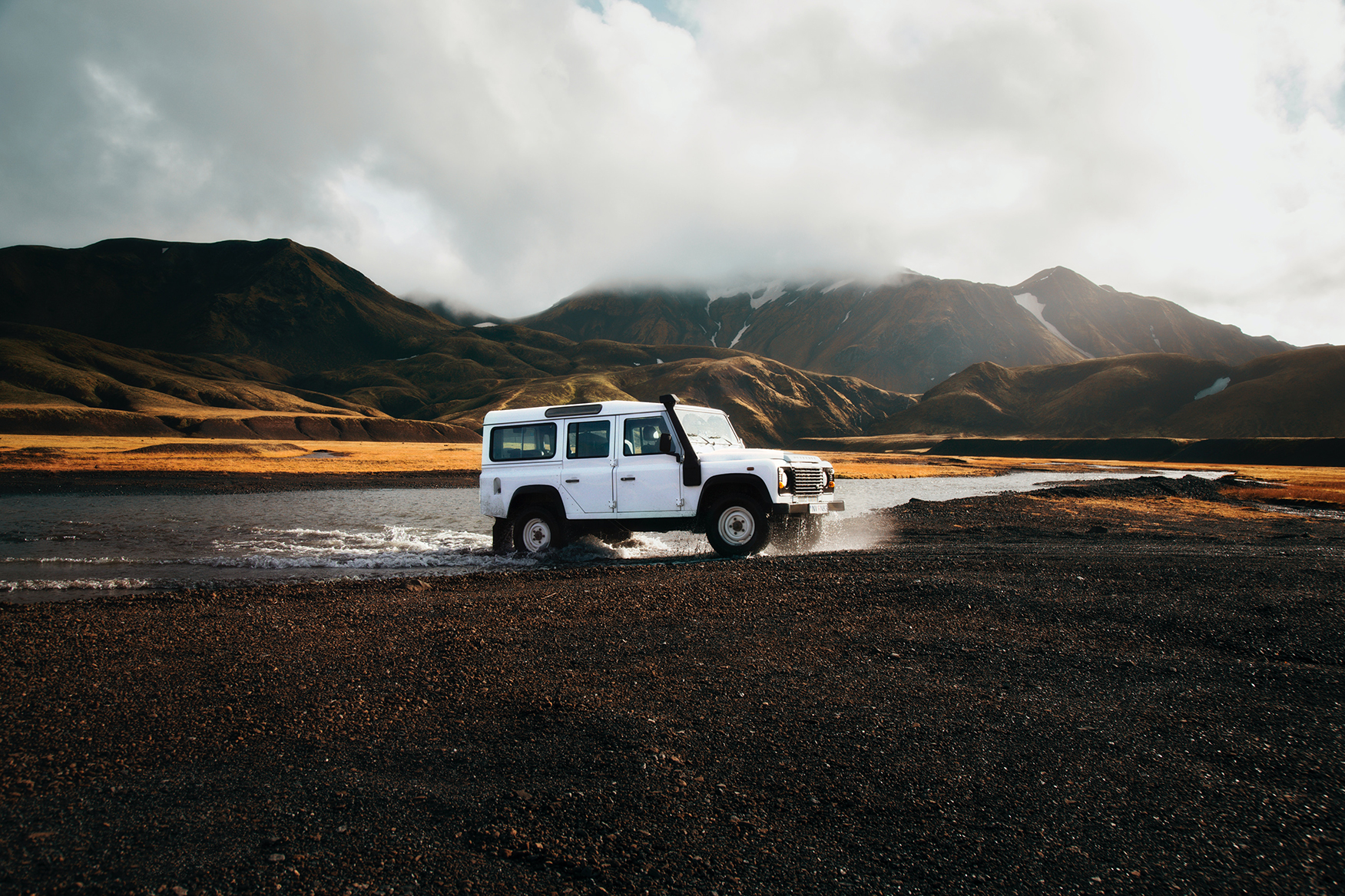 A white SUV exiting water onto black sand with the mountains of  Landmannalaugar, Iceland in the background