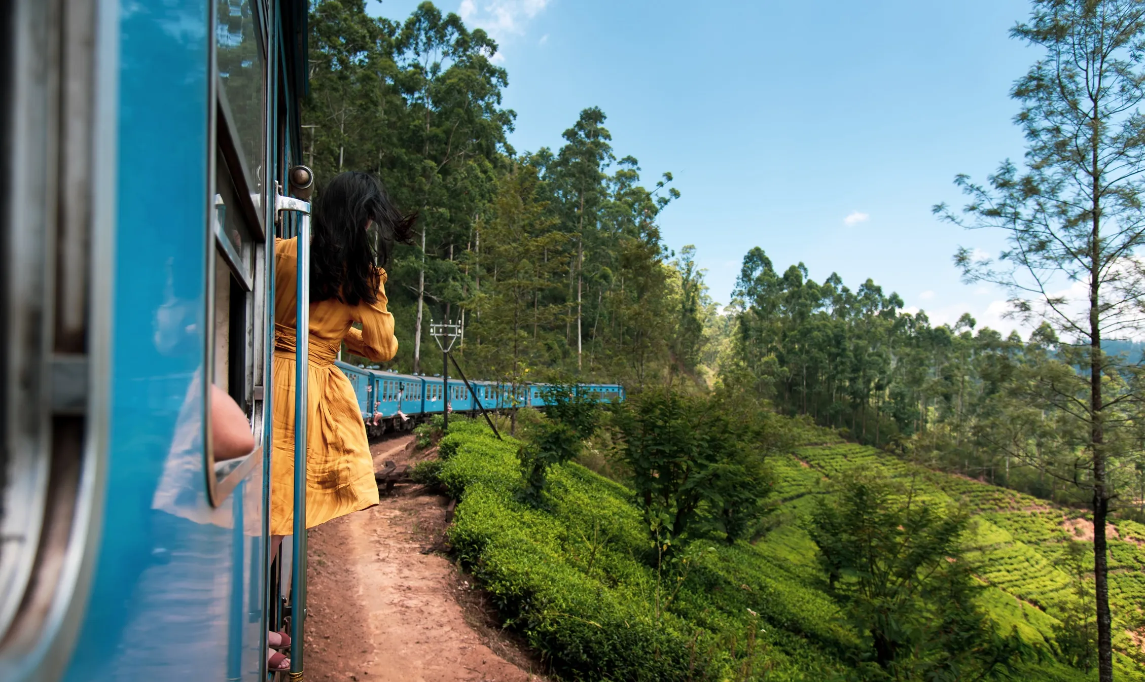 A woman in a yellow dress leans out of a blue train traveling through a lush green landscape with trees and hills.